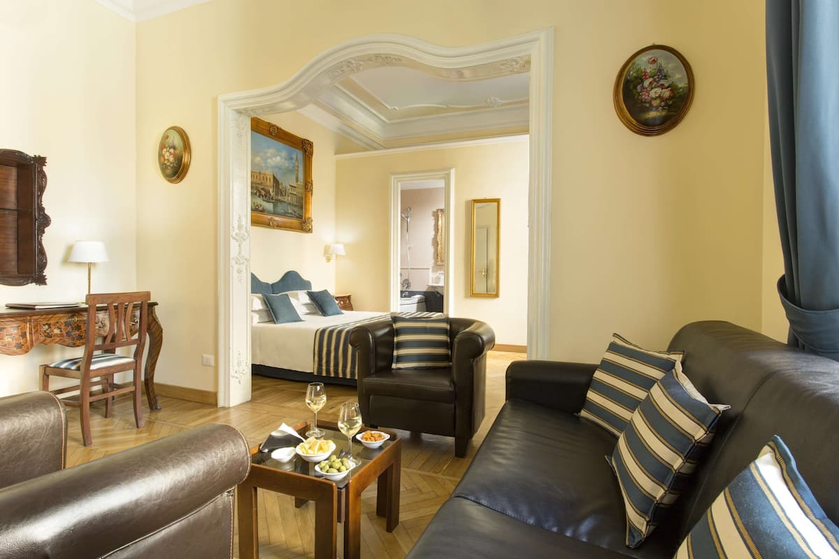 A living room with a sofa, armchair, dining table, and a bed in the background.