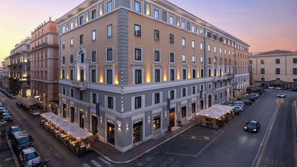 Un hôtel à plusieurs étages, doté d’une architecture classique, avec un rez-de-chaussée comprenant un espace extérieur aménagé en terrasse et une série de boutiques.