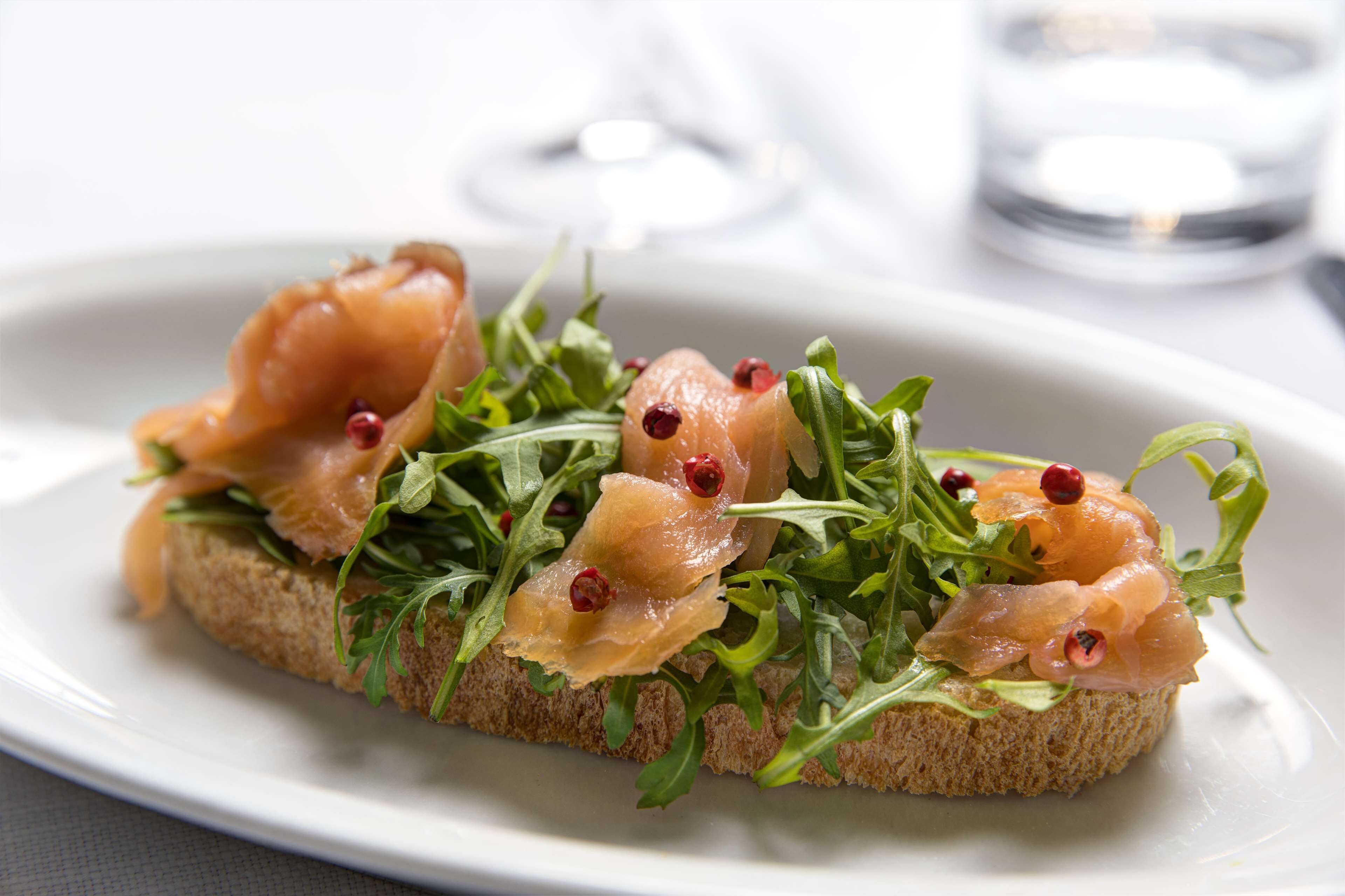 A plate with a sandwich topped with smoked salmon and arugula, accompanied by a glass of water.