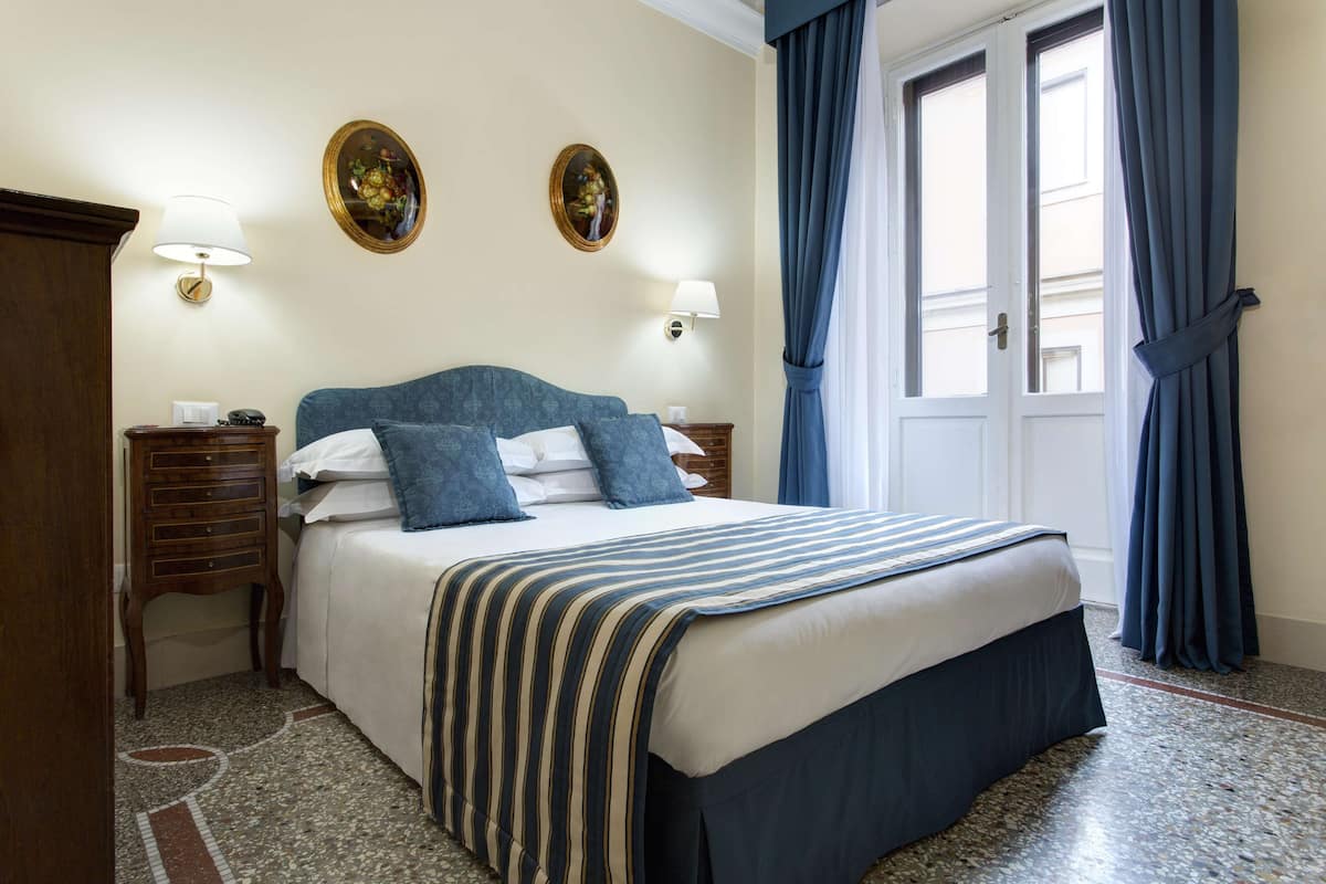 A hotel room with a bed, two bedside tables, a nightstand, a mirror, and a window with curtains.