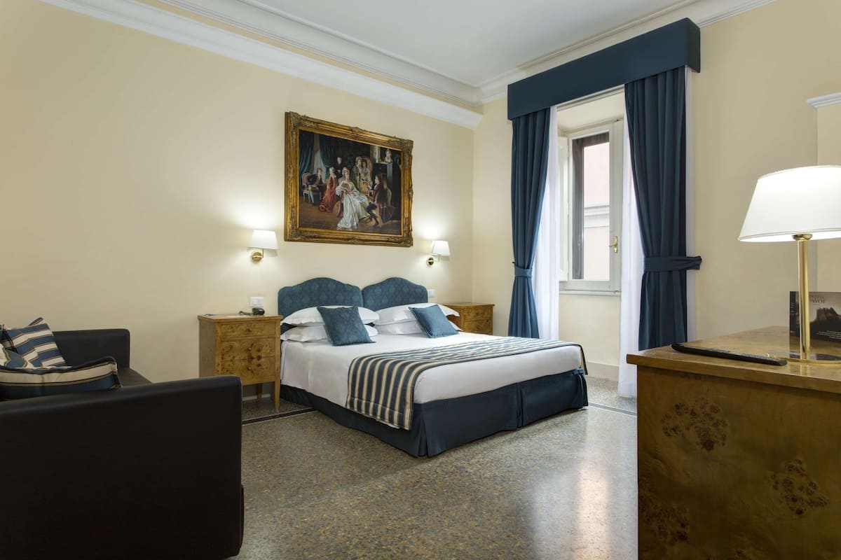 A hotel room with a bed, a nightstand, a chair, a desk, a lamp, a painting, and a window with curtains.