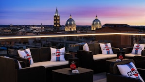 Une terrasse sur le toit, dotée de meubles en osier, de coussins à l’effigie de la marque Martini, et offrant une vue sur les bâtiments et les dômes de la ville au crépuscule.