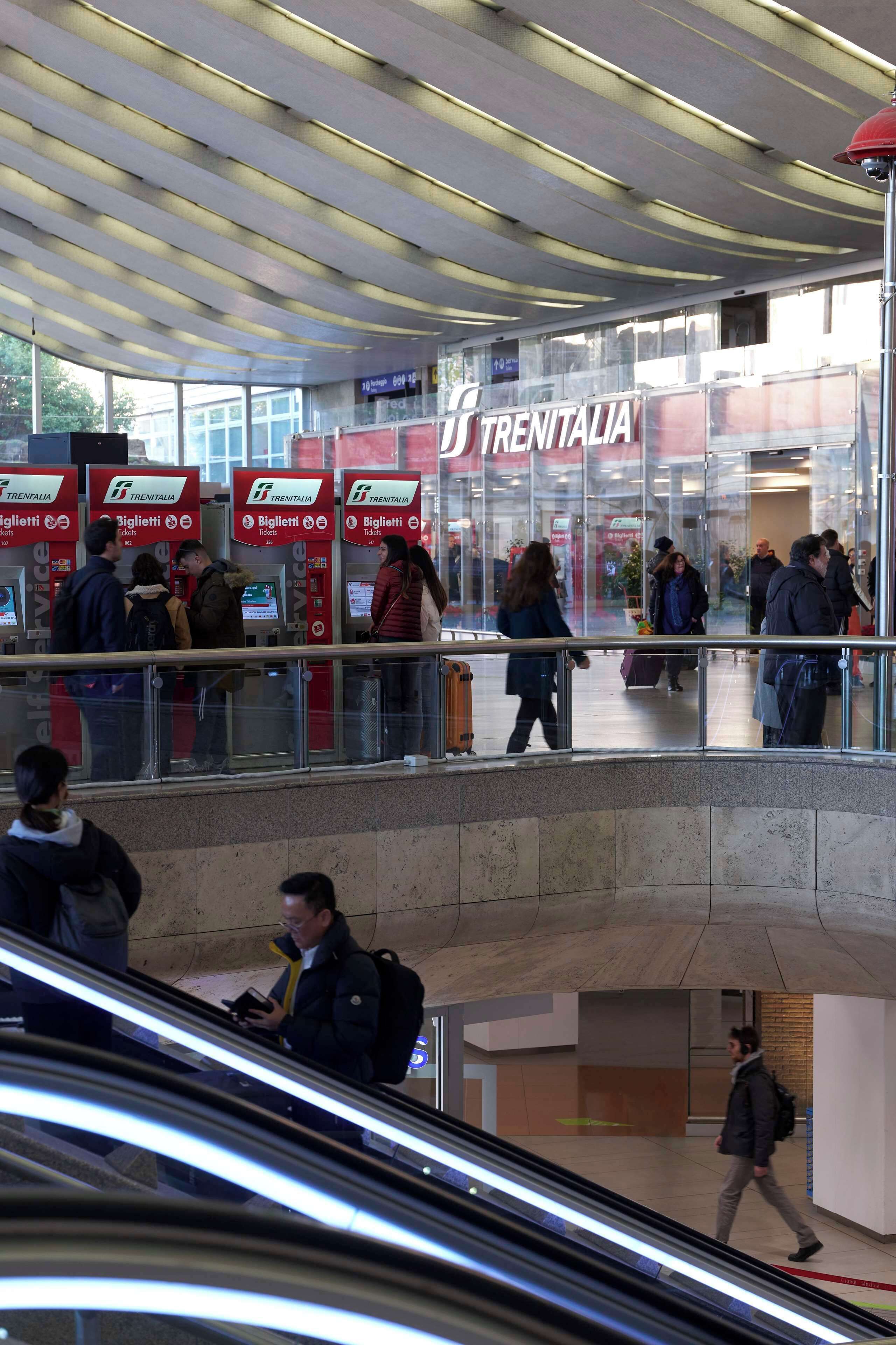 A public transportation hub with multiple signs for Trenitalia, a glass facade, and people using the escalator.