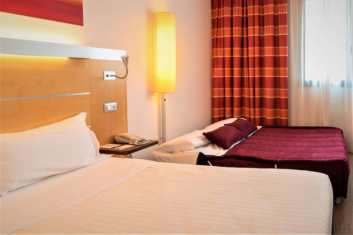 A hotel room with two beds, a bedside table with a lamp, a telephone, and a window with curtains.