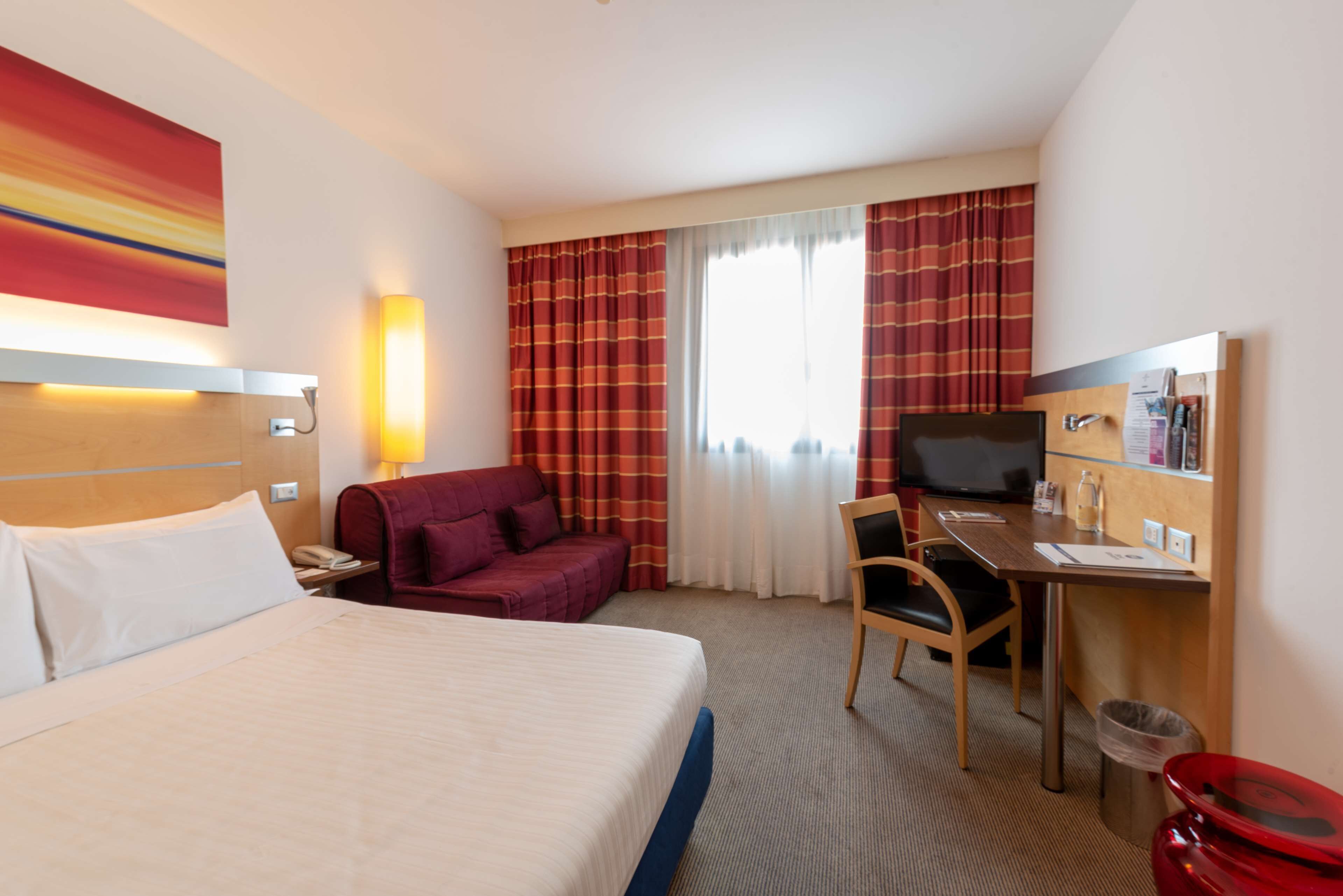 Ein Hotelzimmer mit Bett, Schreibtisch, Stuhl, Fernseher und einem Fenster mit Vorhängen.