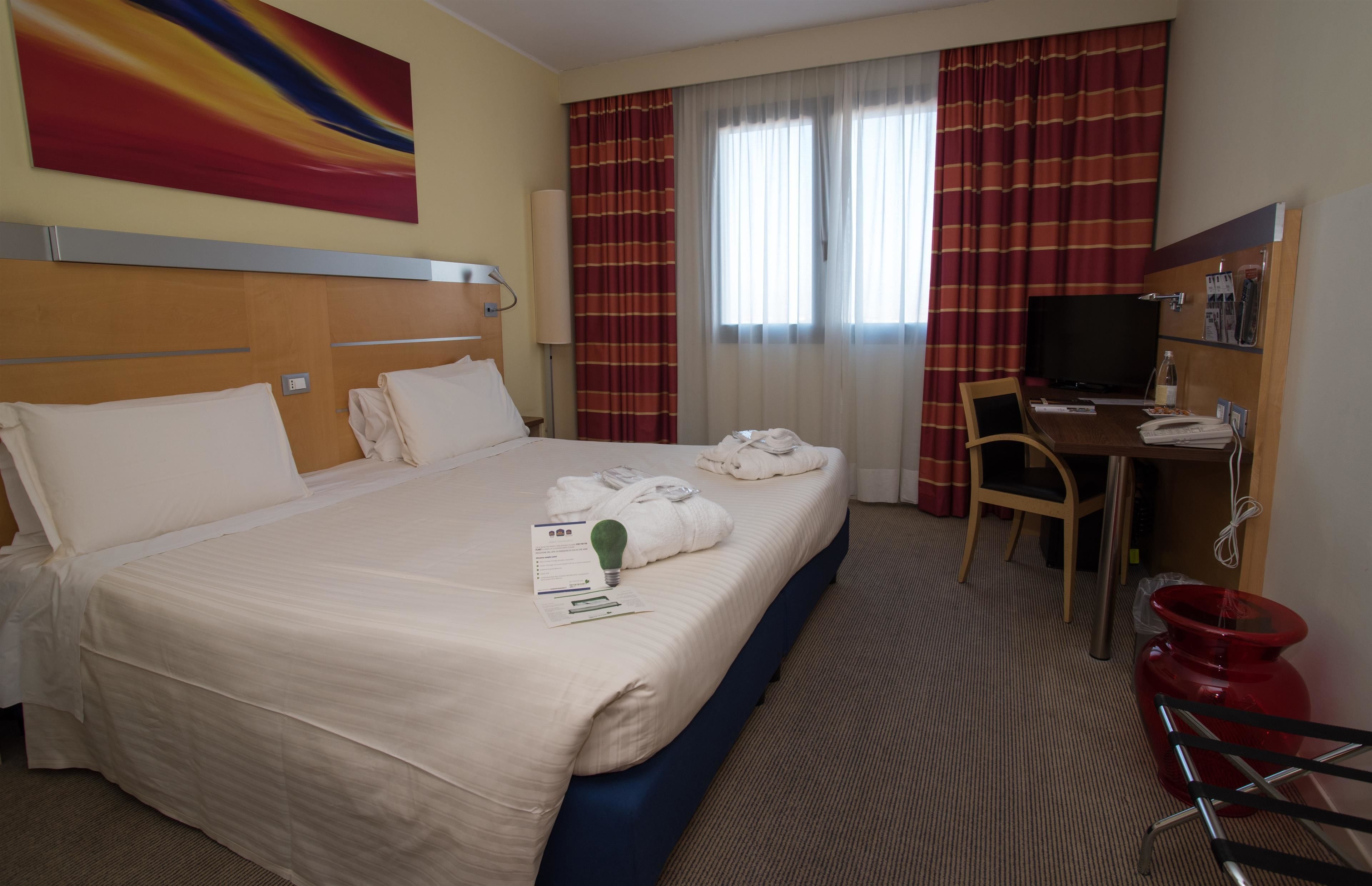 Une chambre d’hôtel avec un grand lit, un bureau avec une chaise, une télévision et une poubelle rouge.