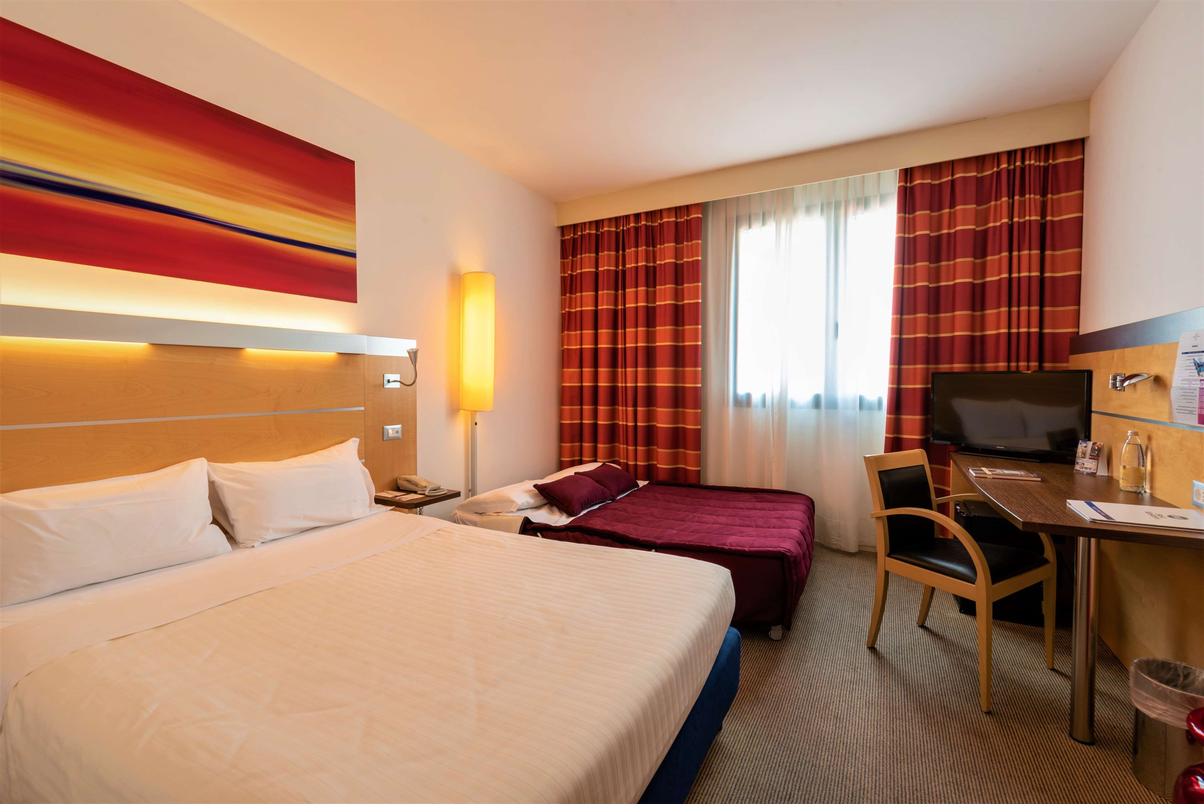Habitación de hotel con una cama grande, un escritorio con silla, televisión y ventana con cortinas.