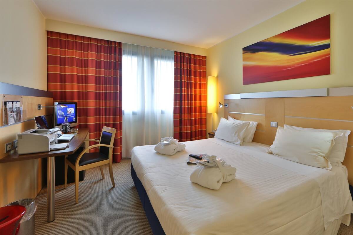 A hotel room with a bed, desk, chair, and a painting on the wall.