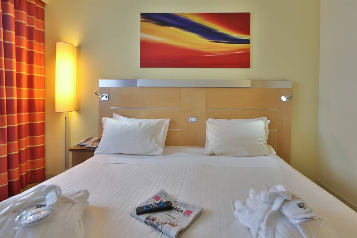 A hotel room with a bed, a painting, a lamp, and a telephone.