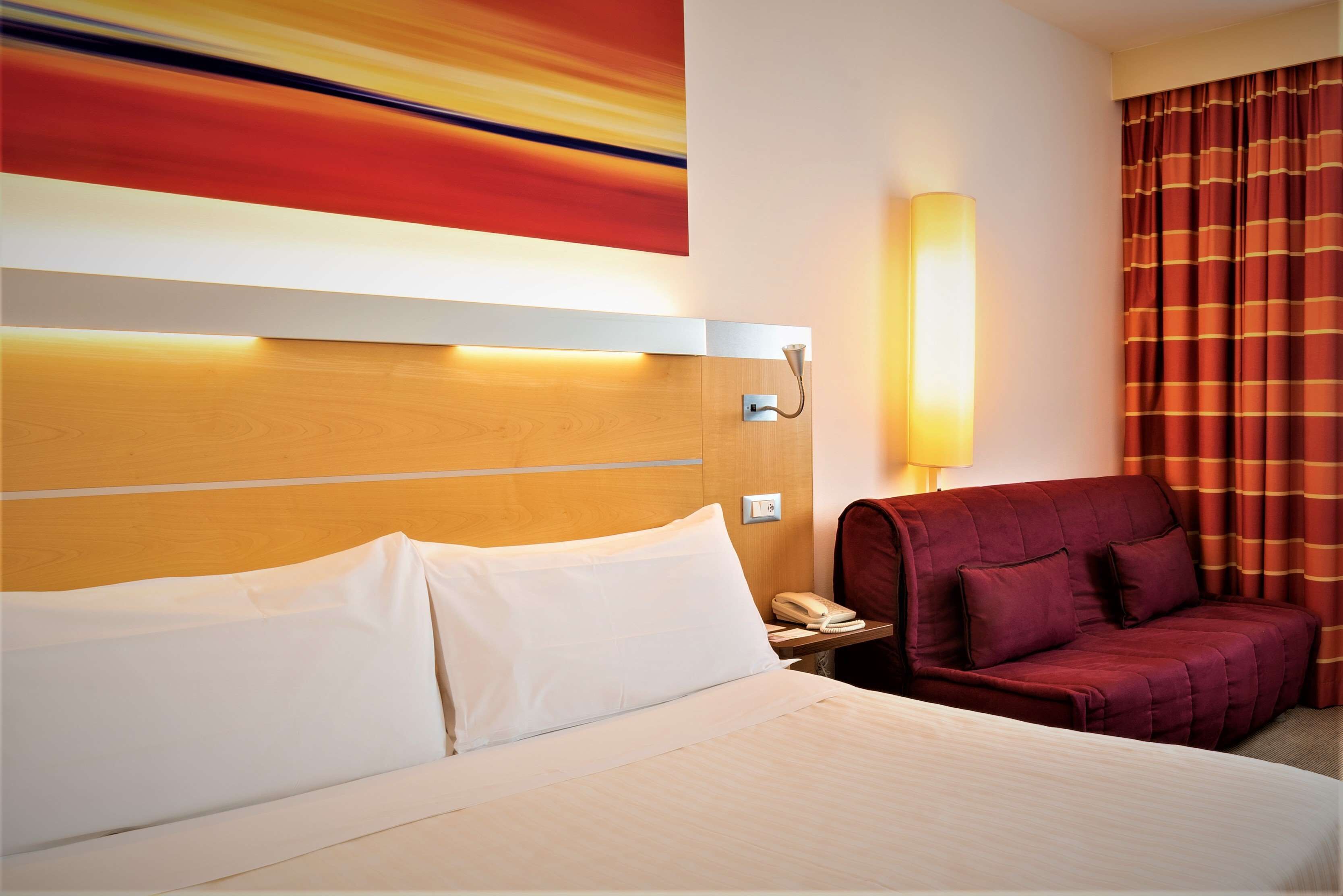 Een hotelkamer met een bed, een rode bank, een nachtkastje met telefoon en een wandlamp.