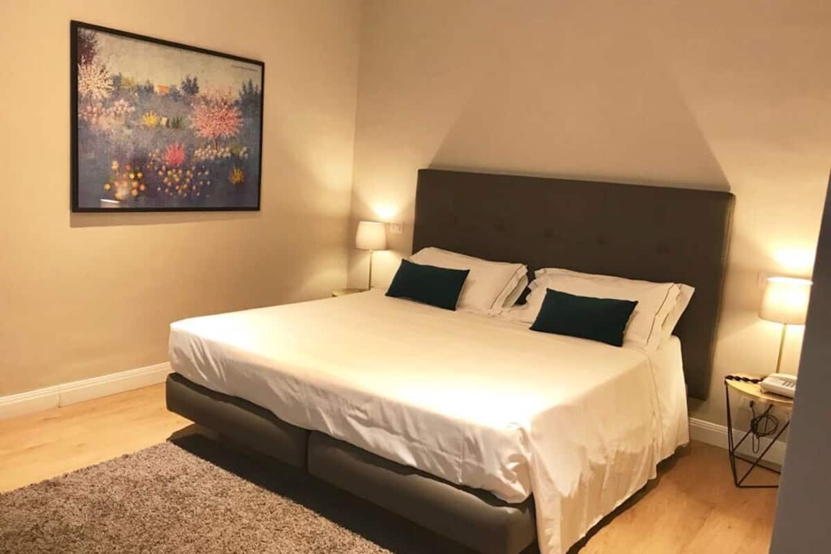 A bedroom with a large bed, two bedside lamps, a framed artwork on the wall, and a carpeted floor.