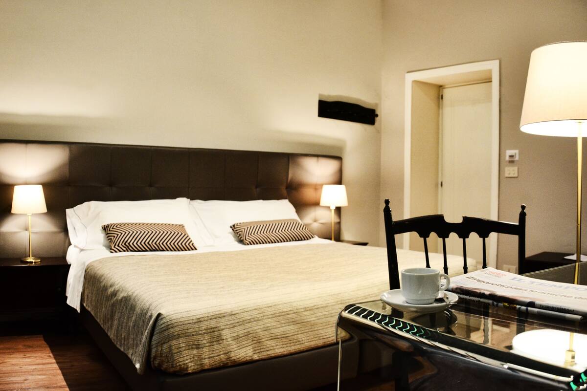 A hotel room with a large bed, two bedside lamps, a wooden chair, a table with a cup and saucer, and a mirror on the wall.