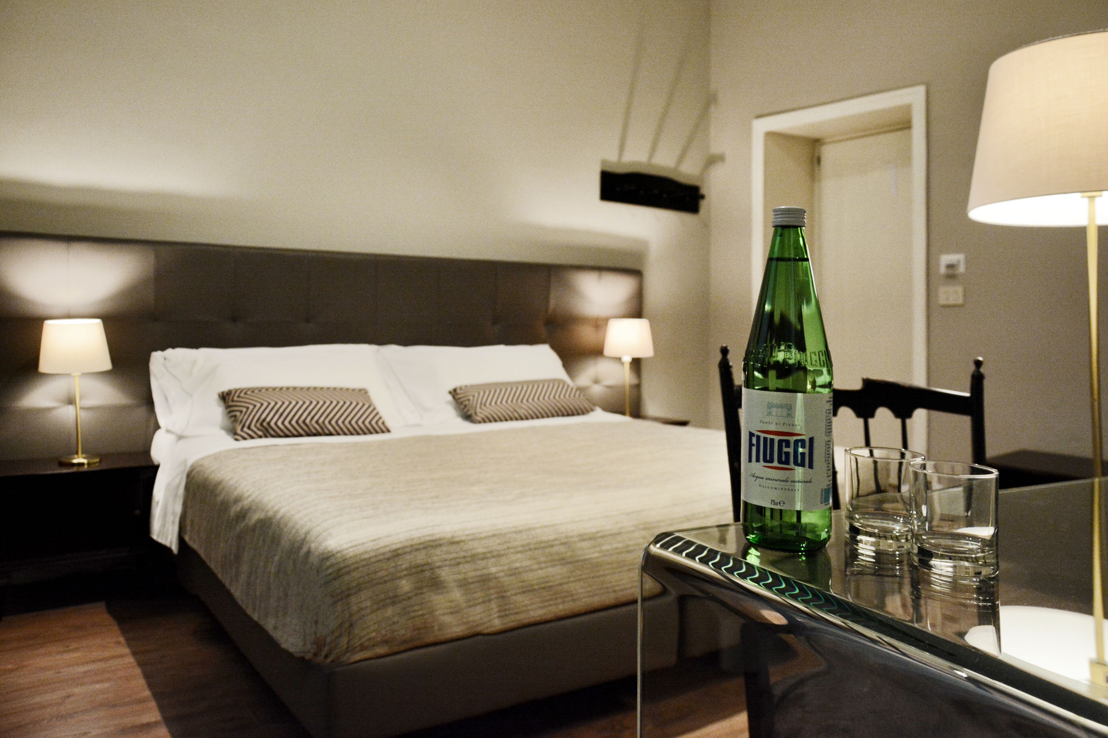 Ein ordentlich bezogenes Bett mit Kopfteil, zwei Kissen, eine grüne Fogg-Flasche auf einem Tisch und zwei Gläser.