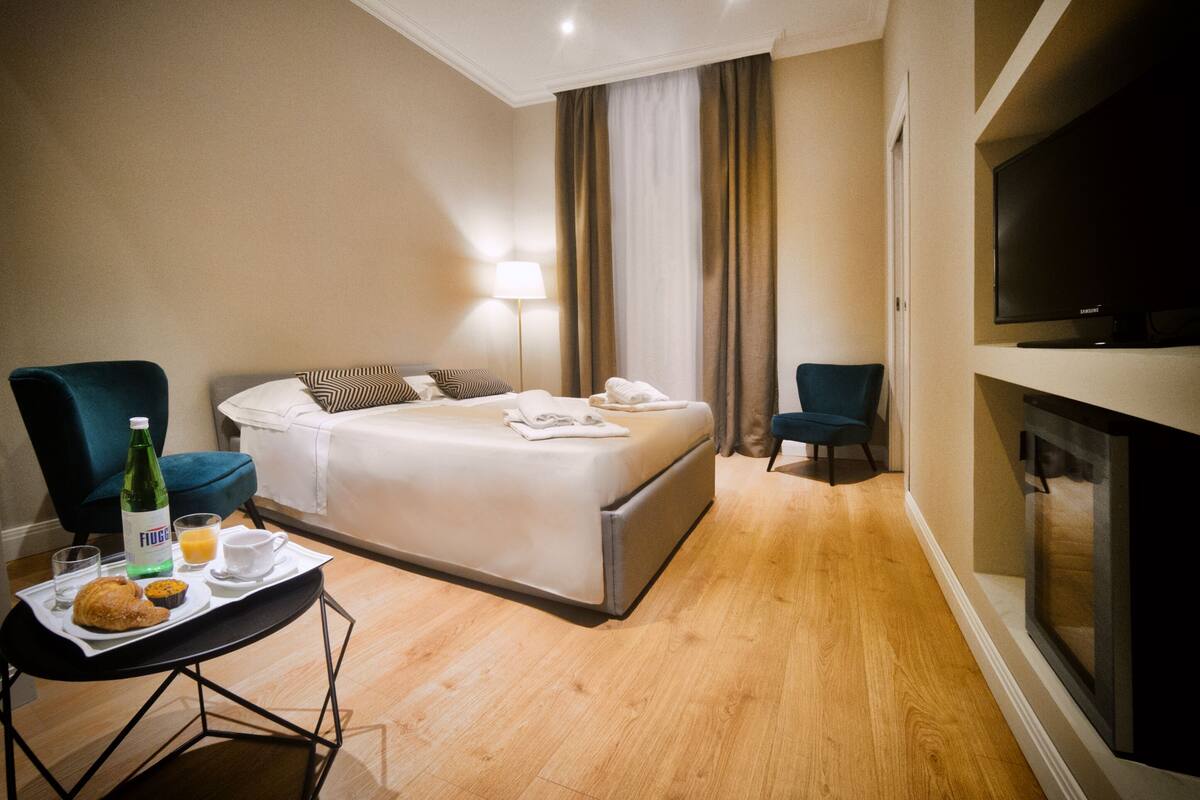 A hotel room with a bed, two armchairs, a TV, a table with breakfast, and a window with curtains.