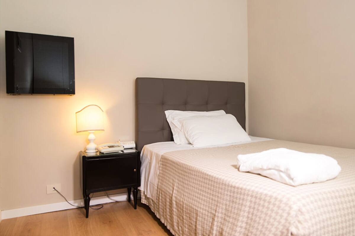 A bedroom with a bed, a nightstand, a lamp, and a television mounted on the wall.