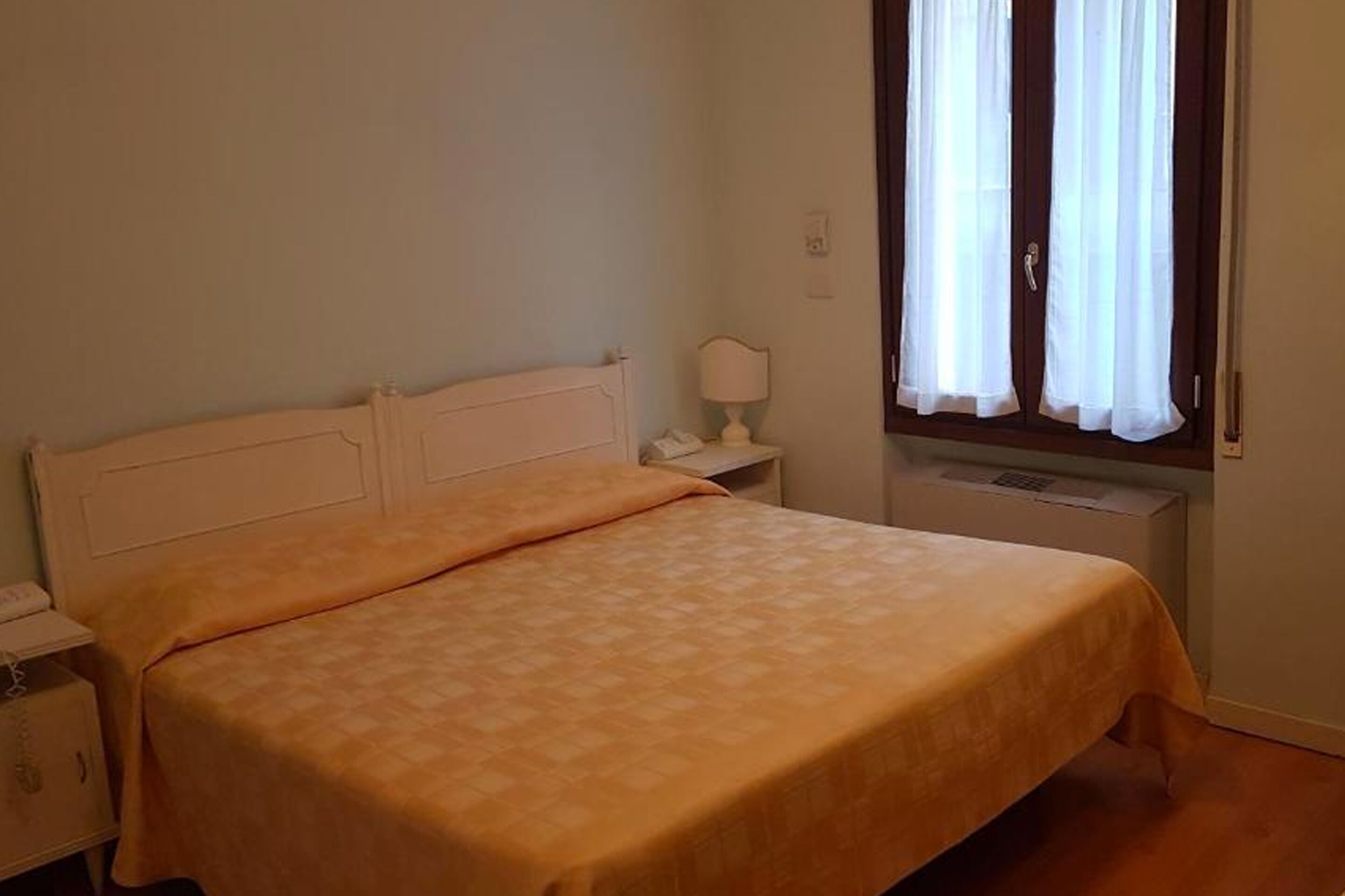 A single bed with a white headboard and a beige bedspread in a room with a window covered by sheer curtains.