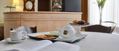 A hotel room with a dining table set for breakfast, including a book, a teapot, a cup, and a plate with pastries.