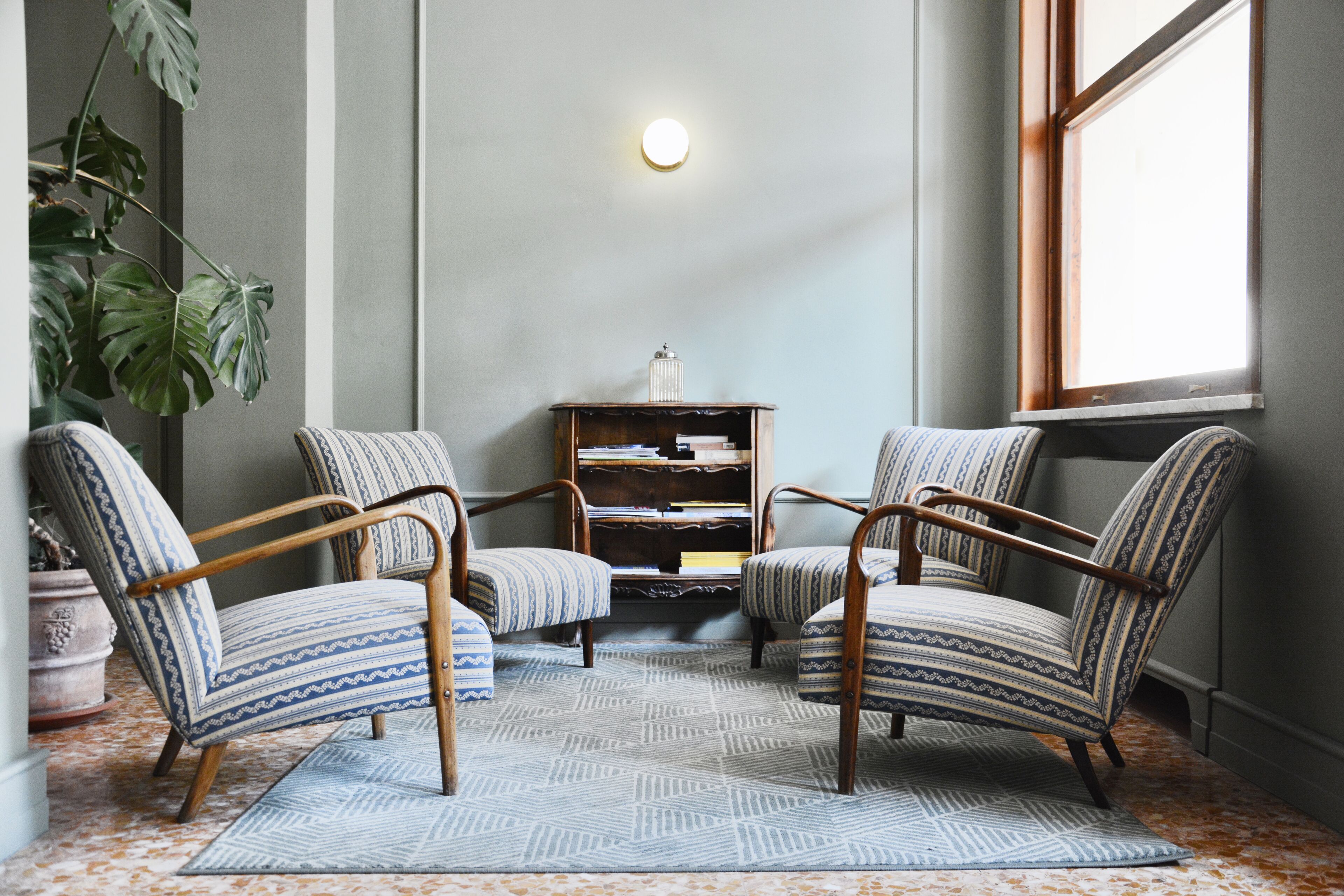 A room with two patterned armchairs, a small wooden side table, a bookshelf, and a large window.