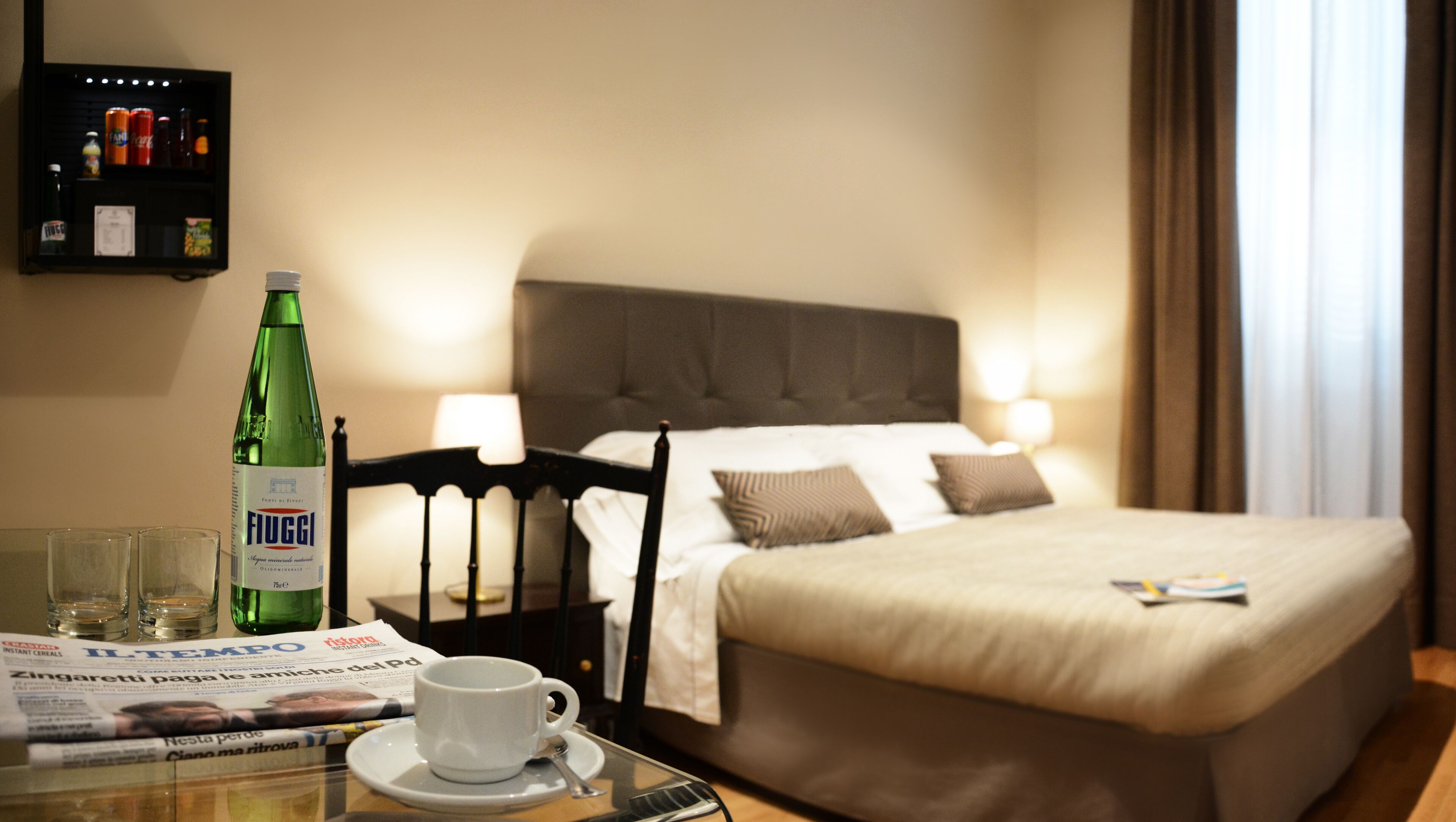 Hotelkamer met een bed, een stoel, een tafel met een kopje en een fles, en een plank met drankjes.