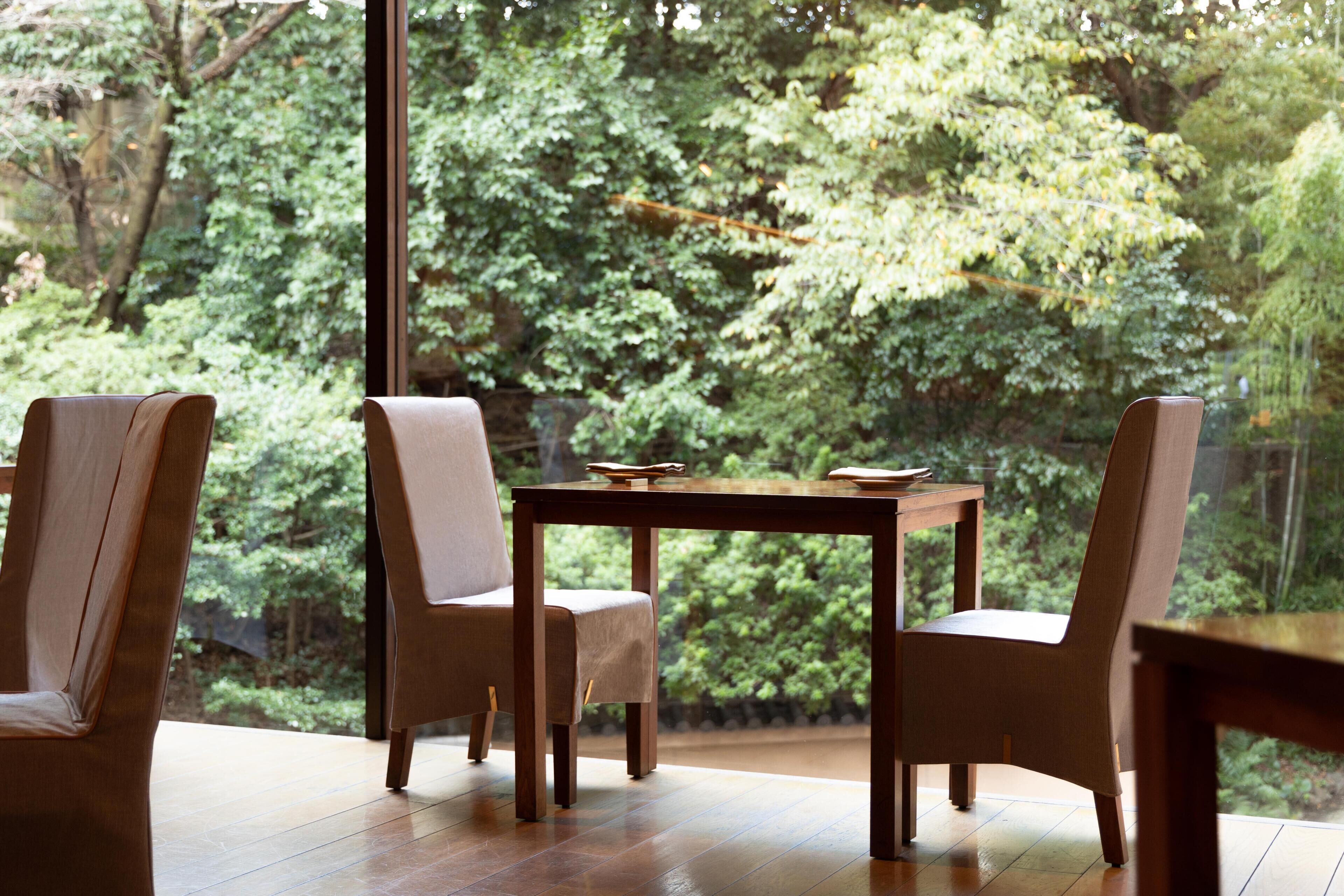 A dining area with a table and chairs, a view of a forest through large windows.
