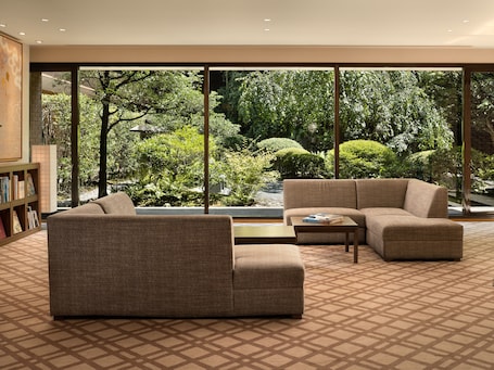 A modern living room with a large window offering a view of a garden. There is a sofa, a coffee table, and a bookshelf.