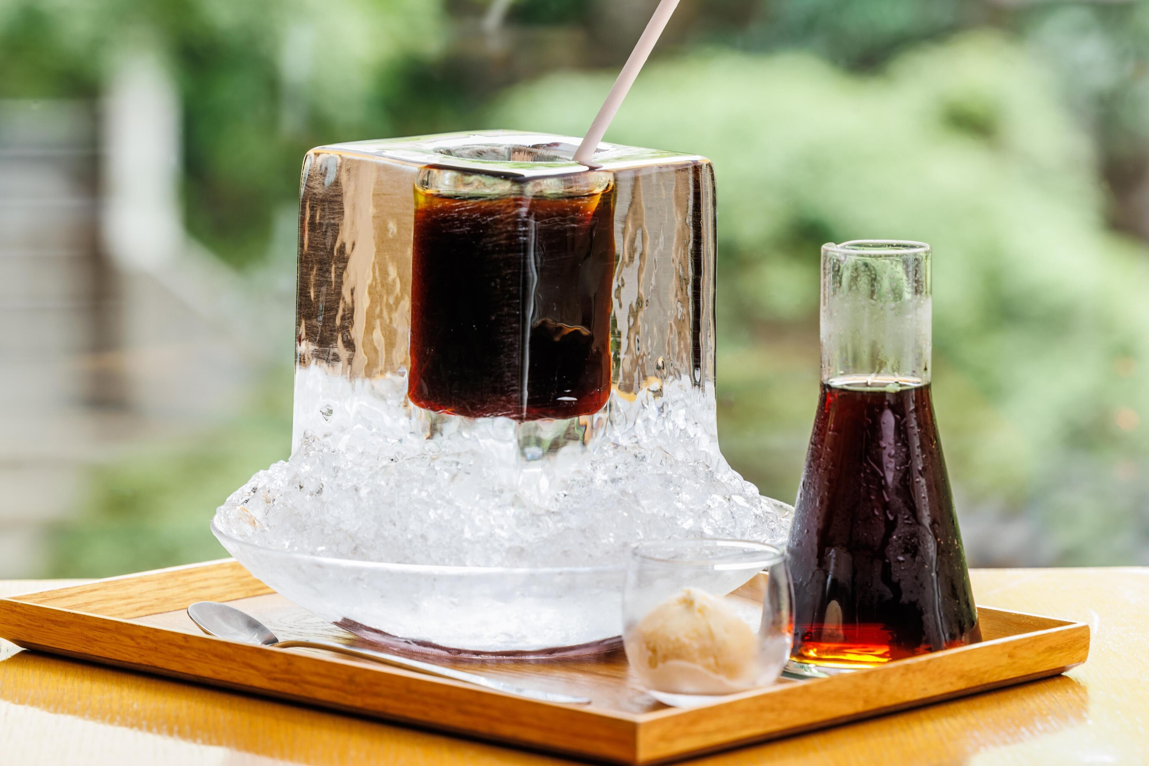A glass of iced tea with a straw, a small glass of syrup, and a spoon on a wooden tray.