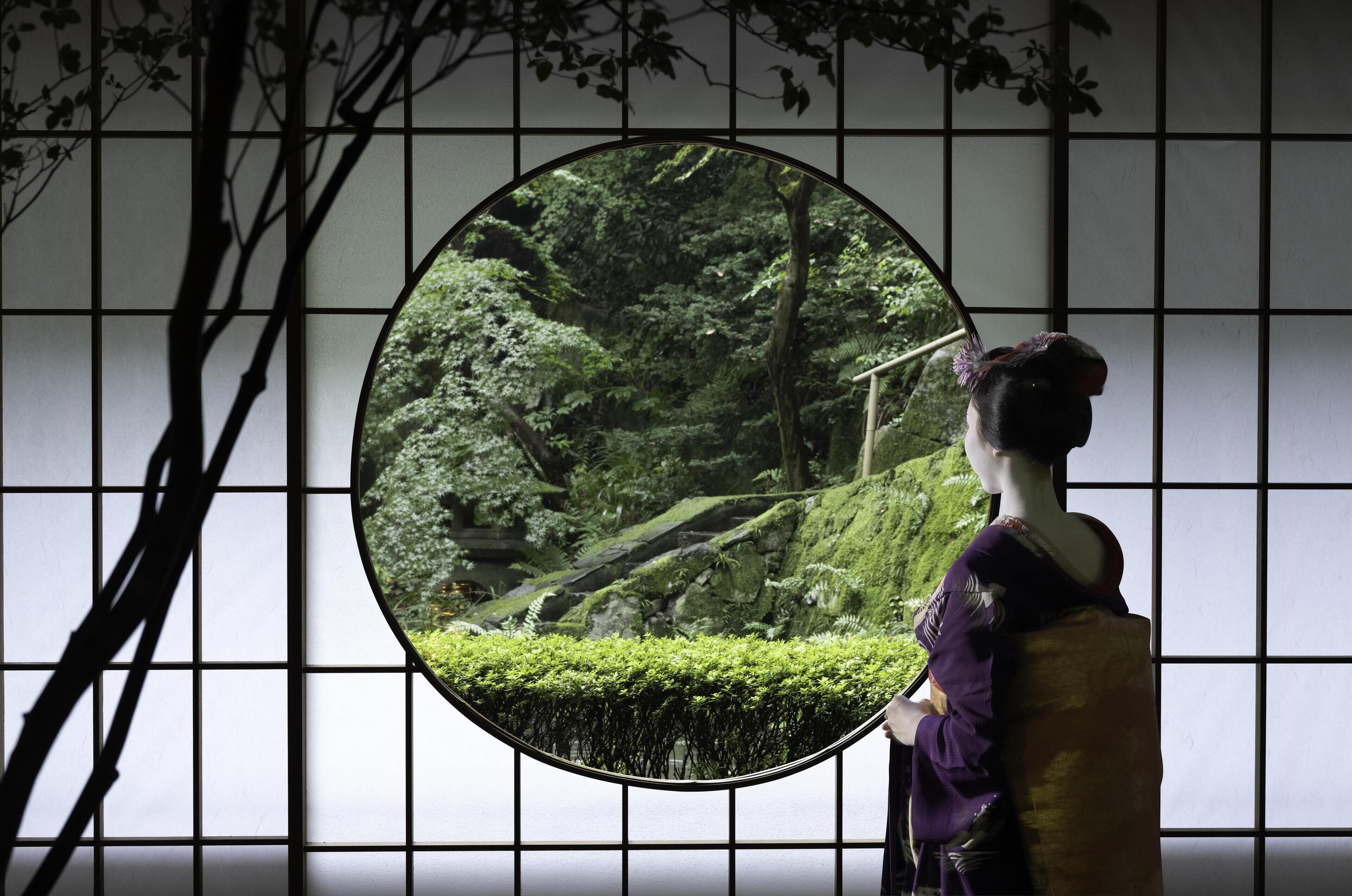 Una habitación japonesa tradicional con suelo de tatami, pantalla shoji y vista a un jardín exuberante a través de un ventanal circular.