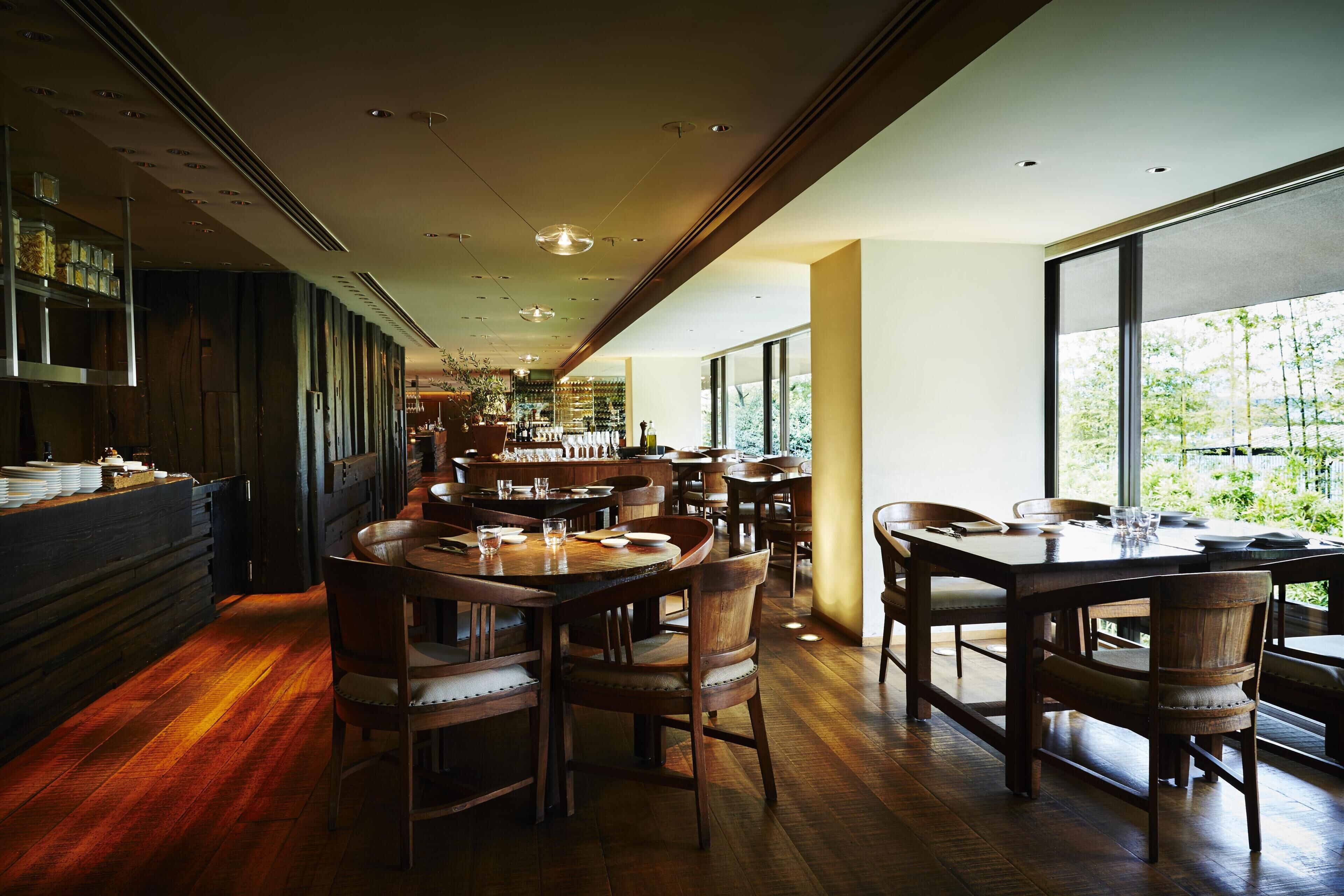 Un restaurante con mesas y sillas de madera, una zona de bar y amplios ventanales con vistas a zonas verdes.