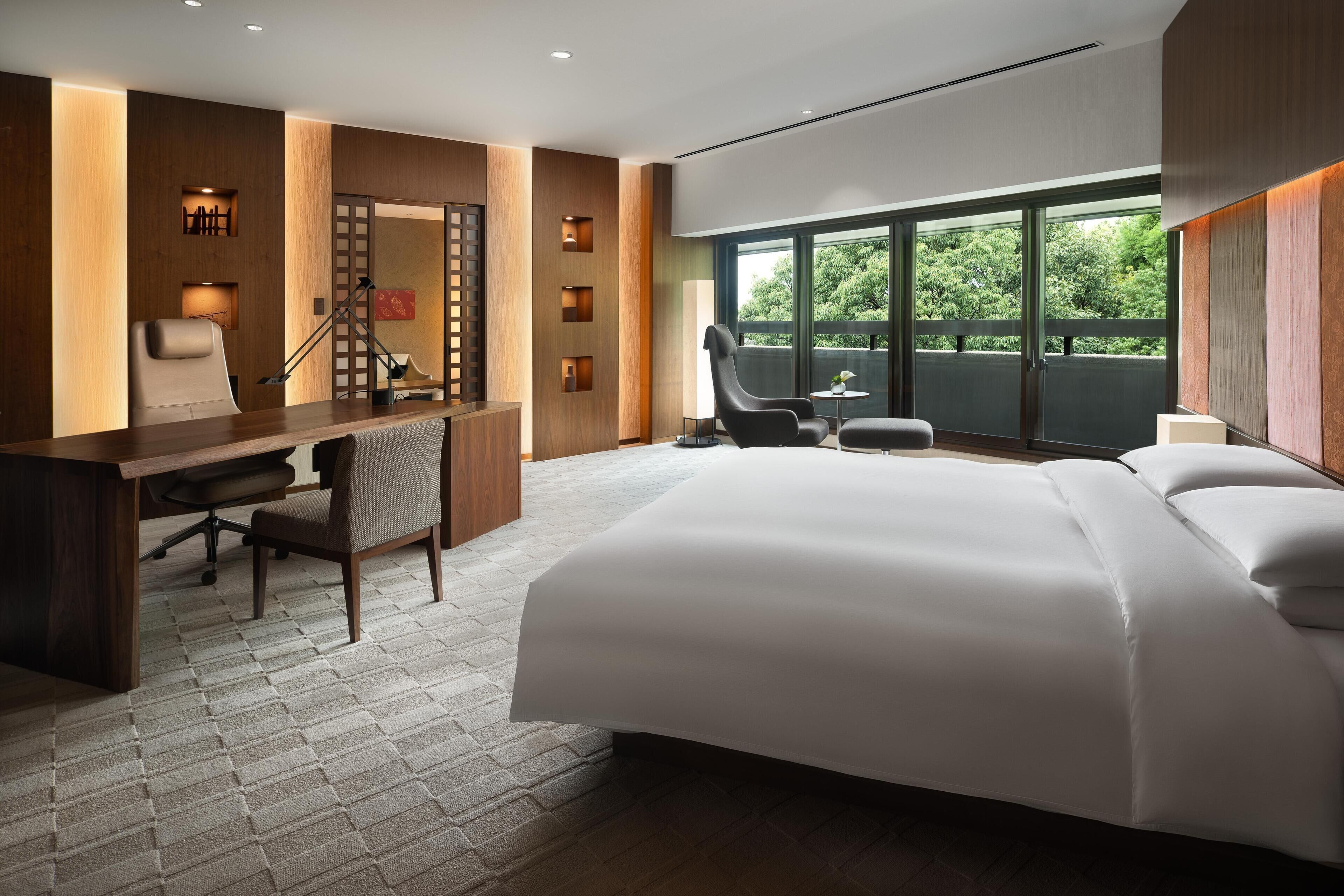 A hotel room with a large bed, a desk, a chair, and a balcony with a view of greenery.