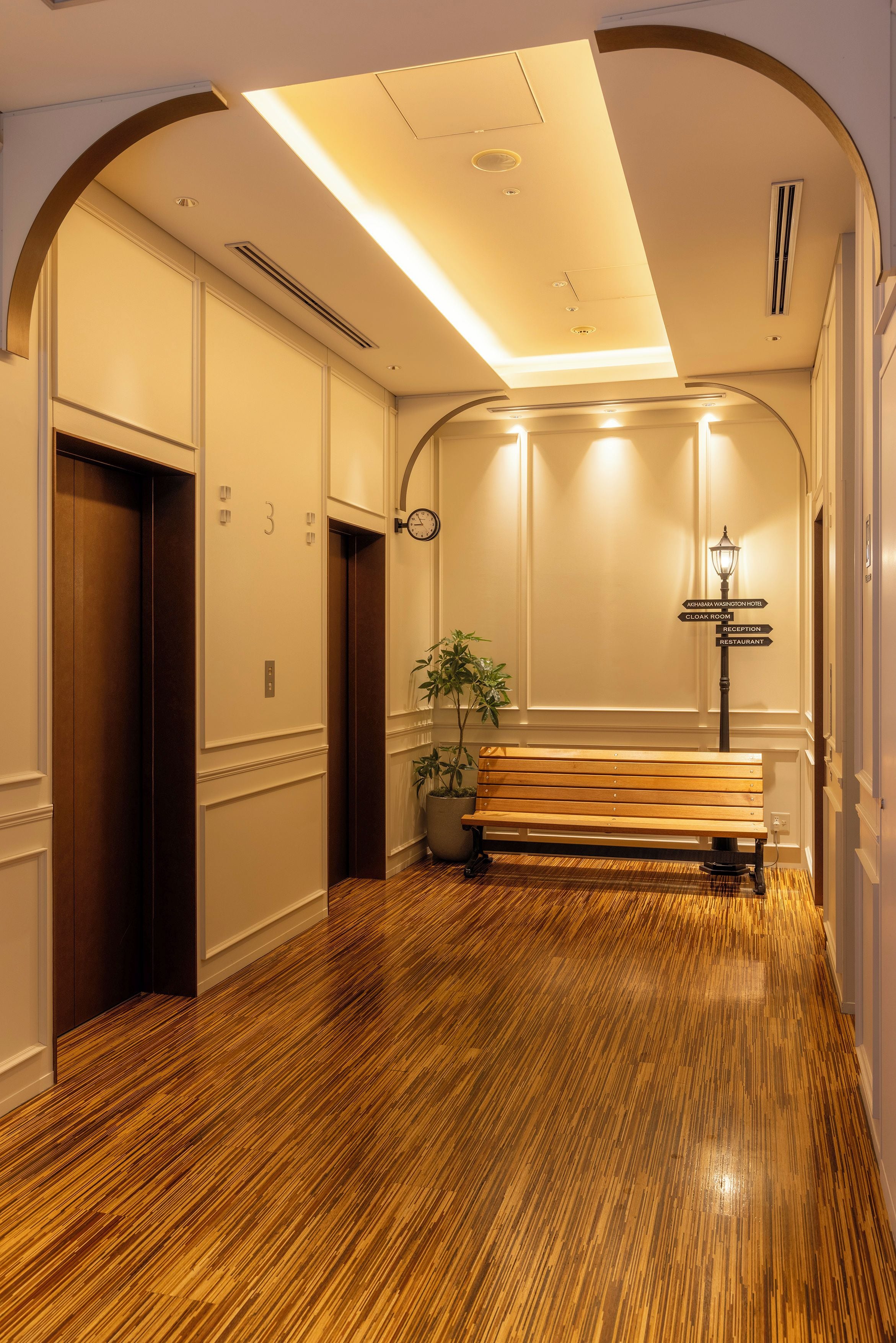 A hotel corridor with wooden flooring, an elevator, a bench, and directional signs to the cloak room, reception, and restaurant.