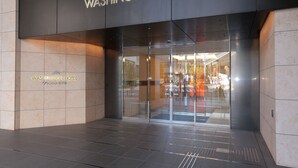 La entrada del Hotel Washington, con fachada moderna, puertas de vidrio y un interior visible a través del cristal.