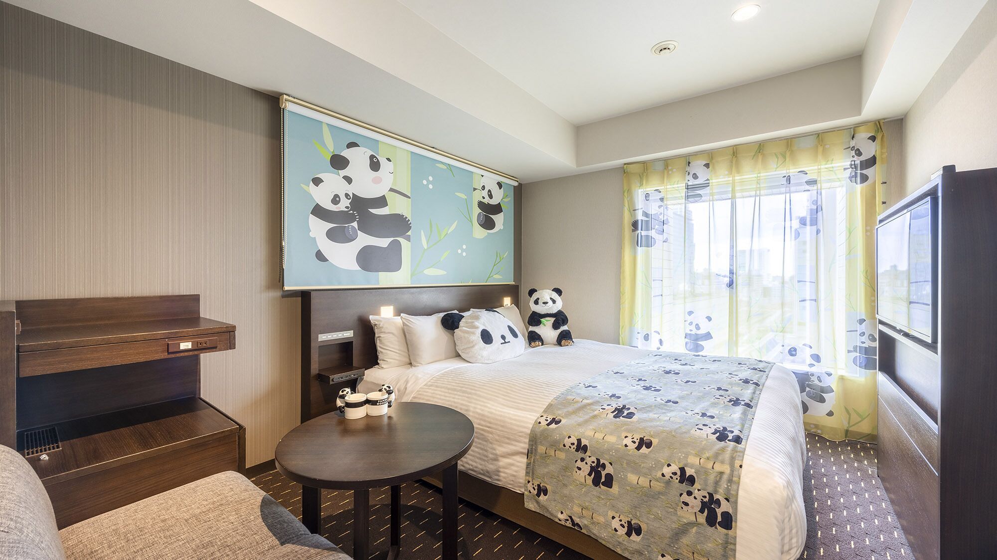 A hotel room with a bed, bedside table, a sofa, a TV, and a panda-themed artwork on the wall.