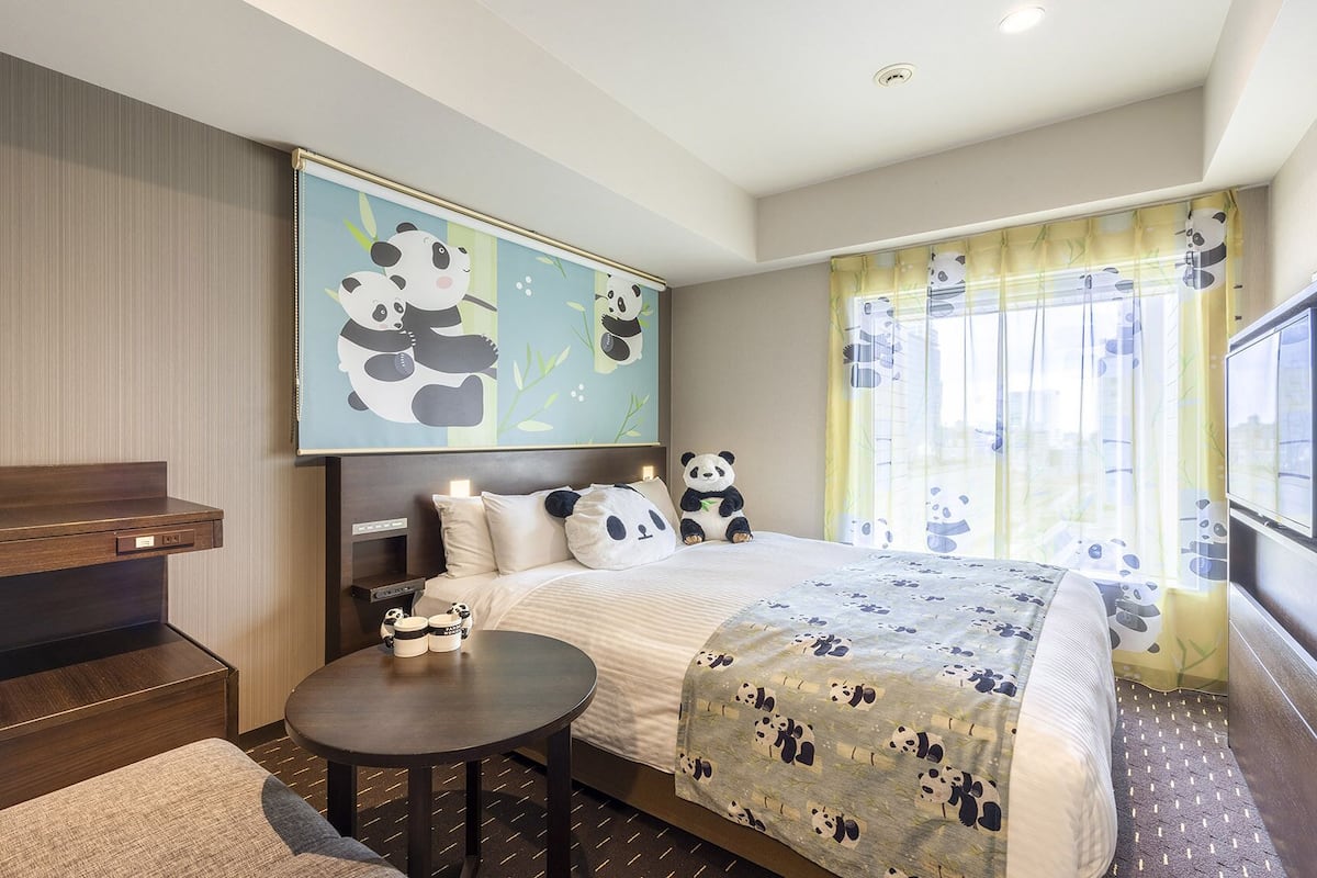 A hotel room with a bed, bedside table, a sofa, a TV, and a panda-themed artwork on the wall.