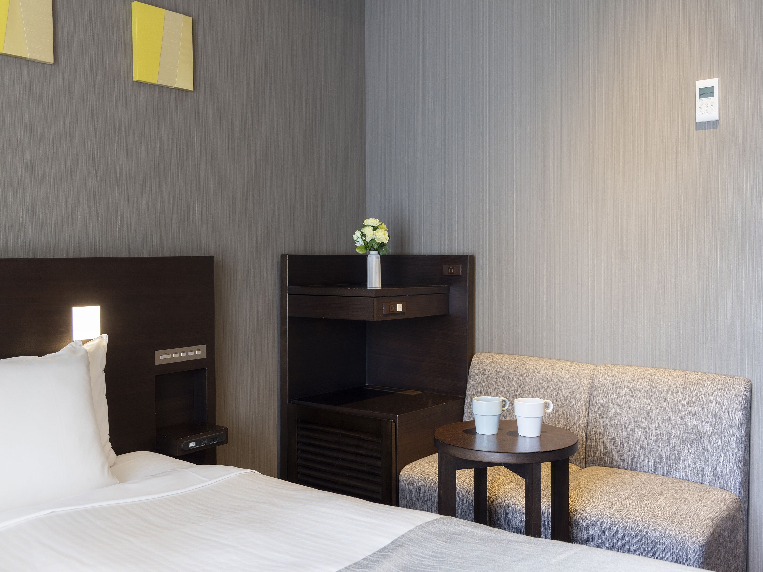 A hotel room with a bed, bedside table, and a small table with two cups.