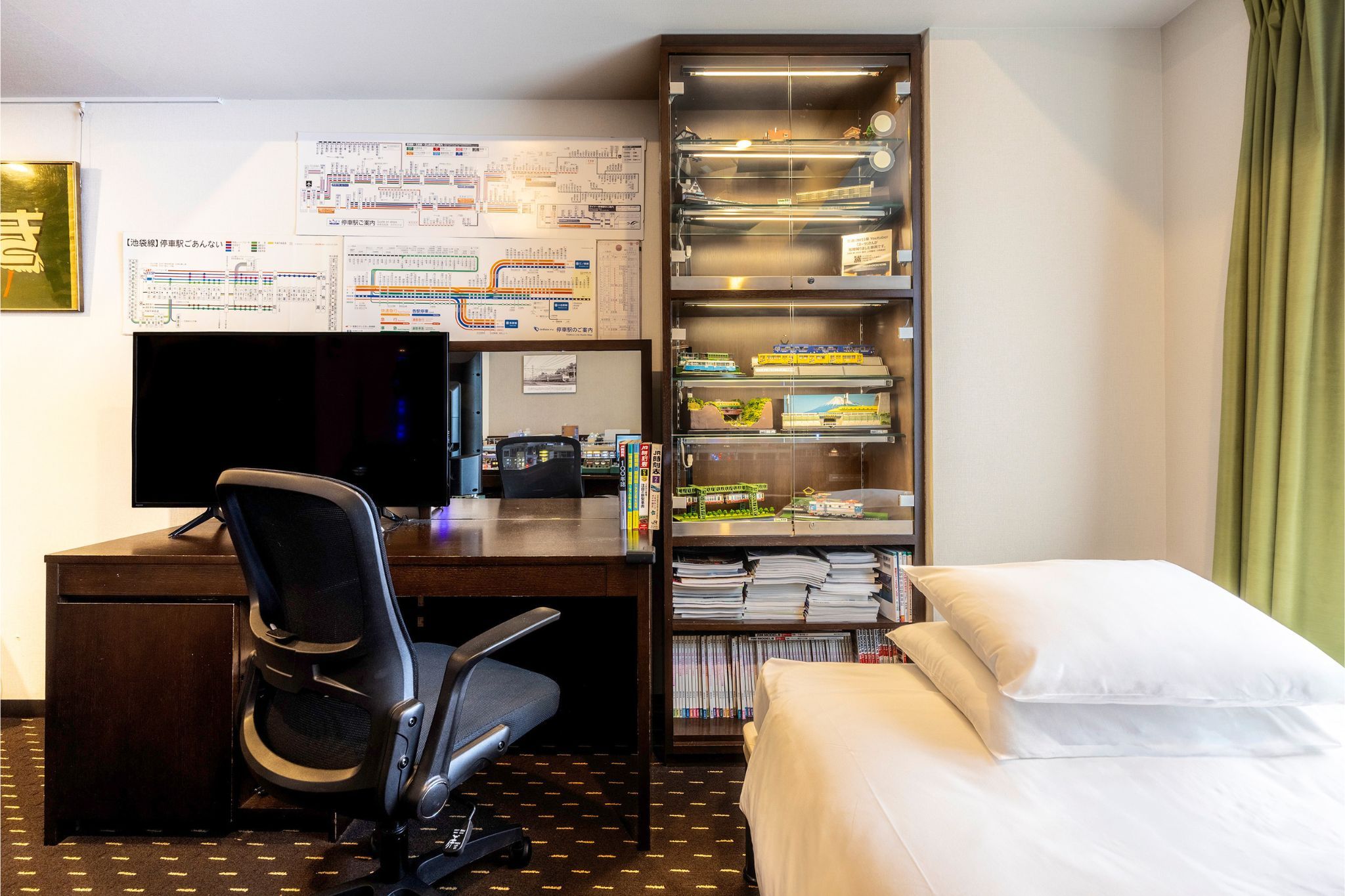 A hotel room with a desk, chair, bed, and a shelf with books and model trains.