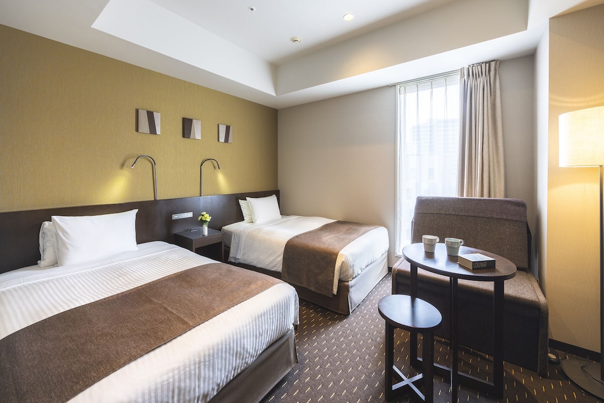 A hotel room with two beds, a desk, a chair, and a window with curtains.