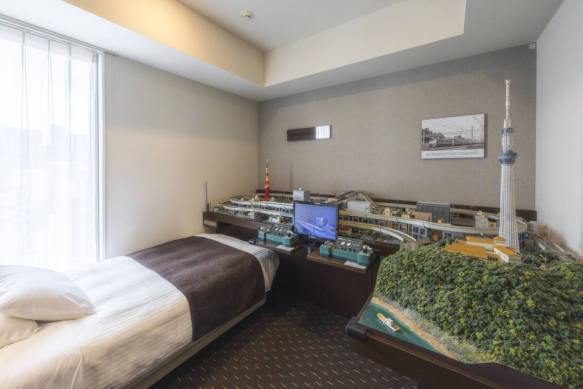 A hotel room with a bed, a desk with a computer, a model train set, and a model of a tower.
