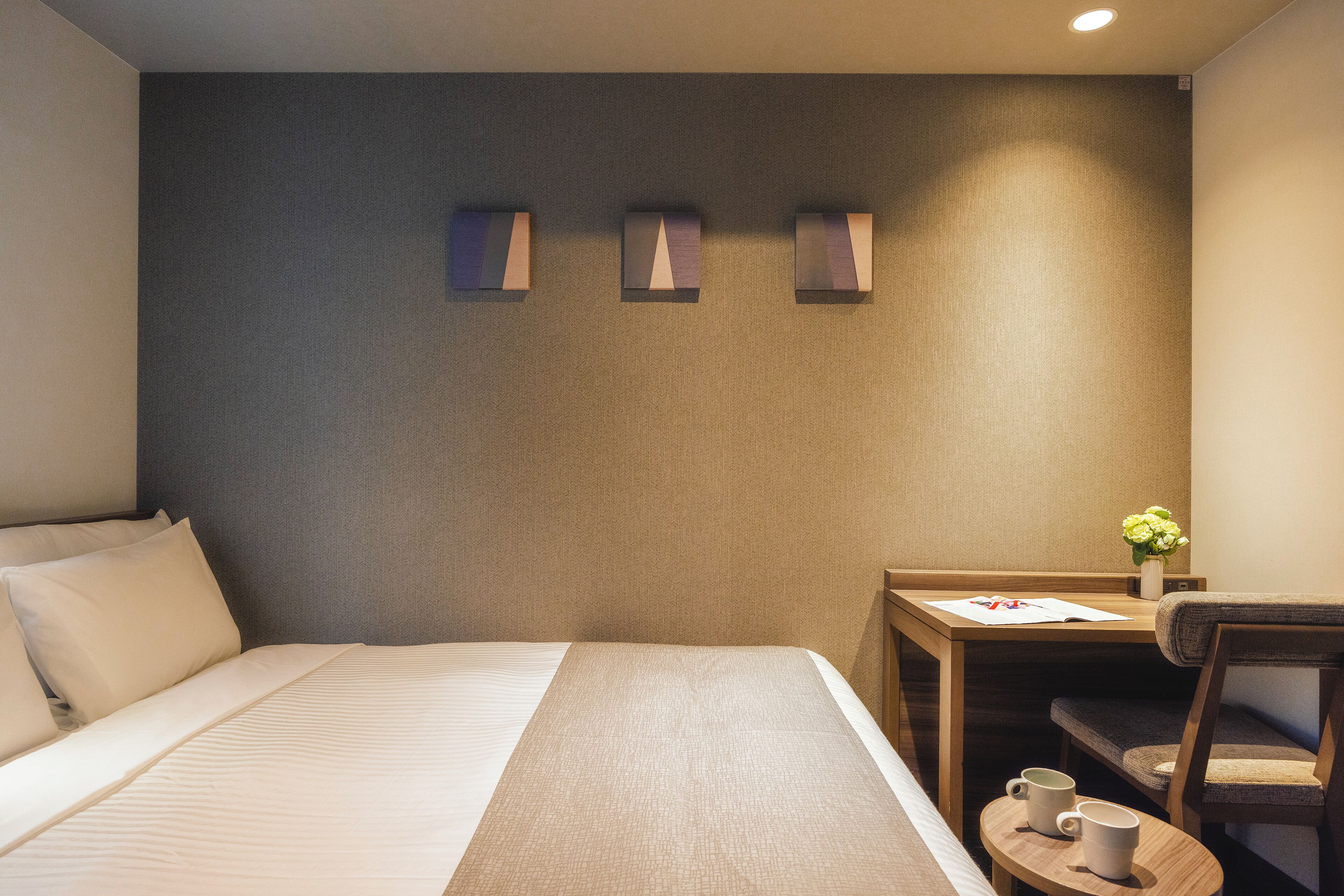 A hotel room with a bed, a desk, a chair, and a small table with two mugs.