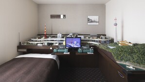Una habitación de hotel con una cama, un escritorio con una computadora, una maqueta de un paisaje urbano y un cuadro en la pared.