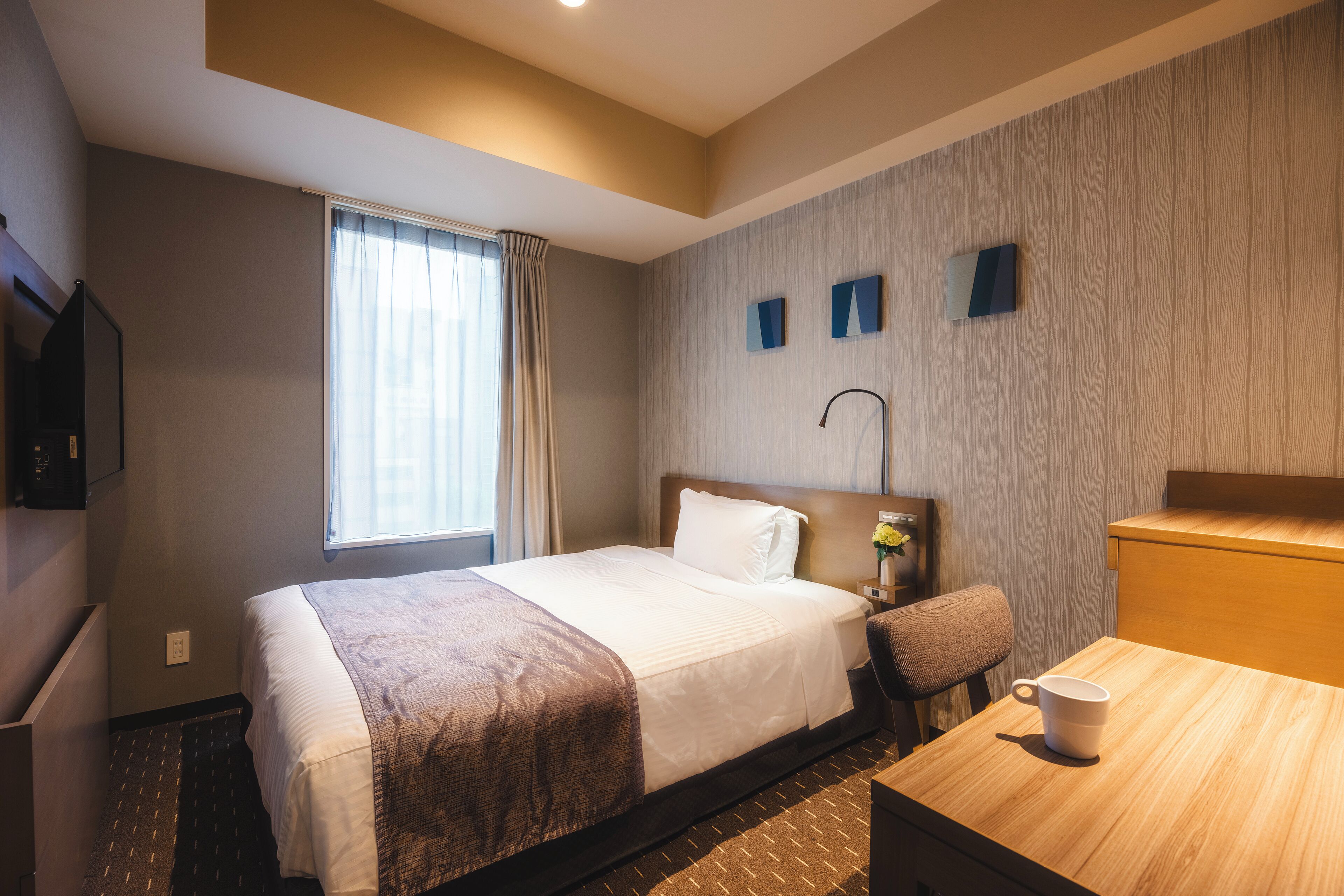 A hotel room with a large bed, a wooden desk, a chair, a TV, and a window with curtains.