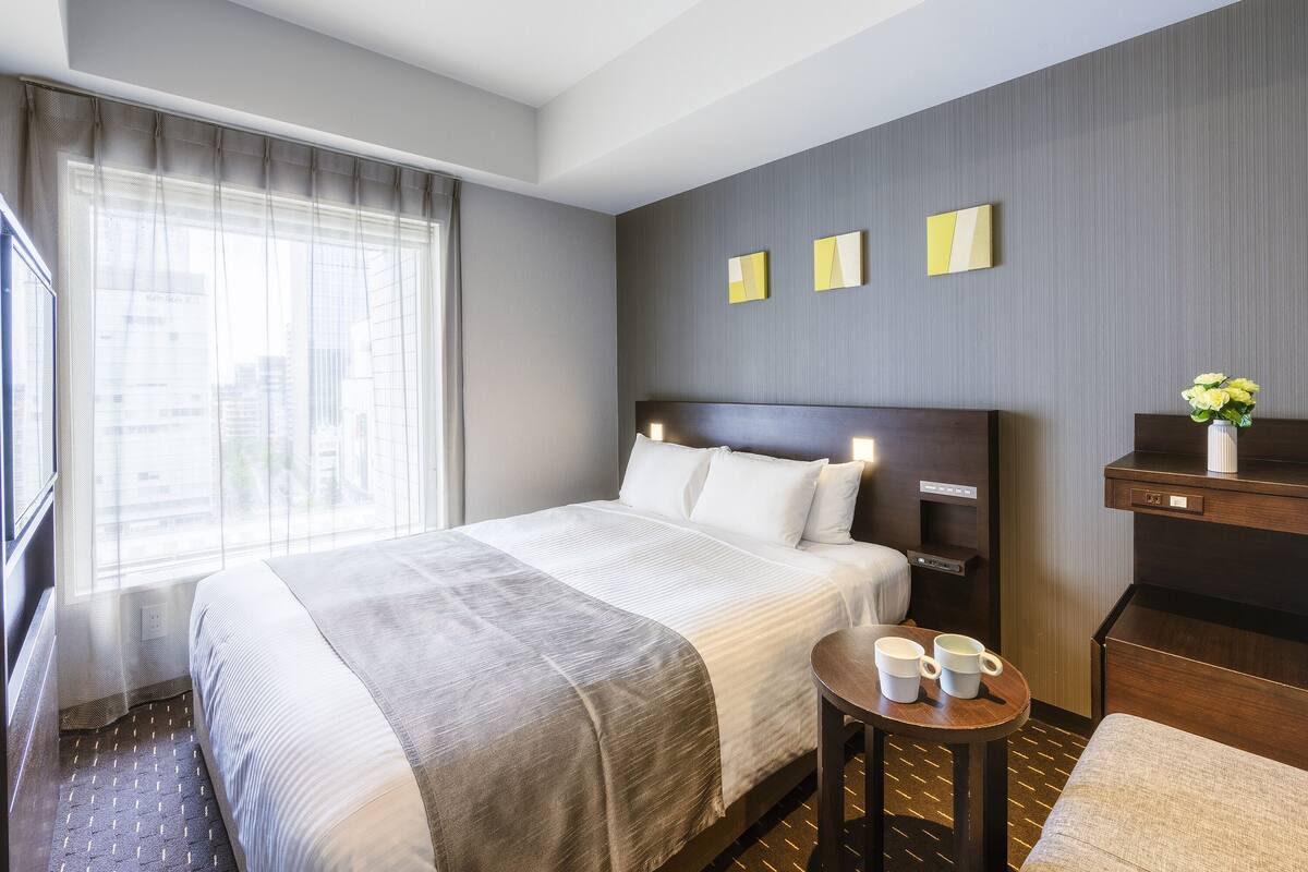 A hotel room with a large bed, a bedside table with two cups, a small desk, and a window with curtains.