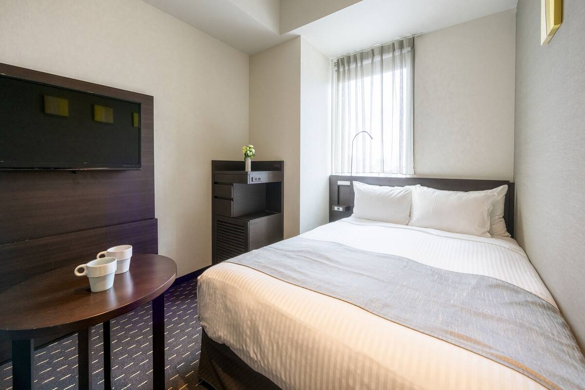 A hotel room with a bed, a TV mounted on the wall, a small table with cups, and a window with curtains.
