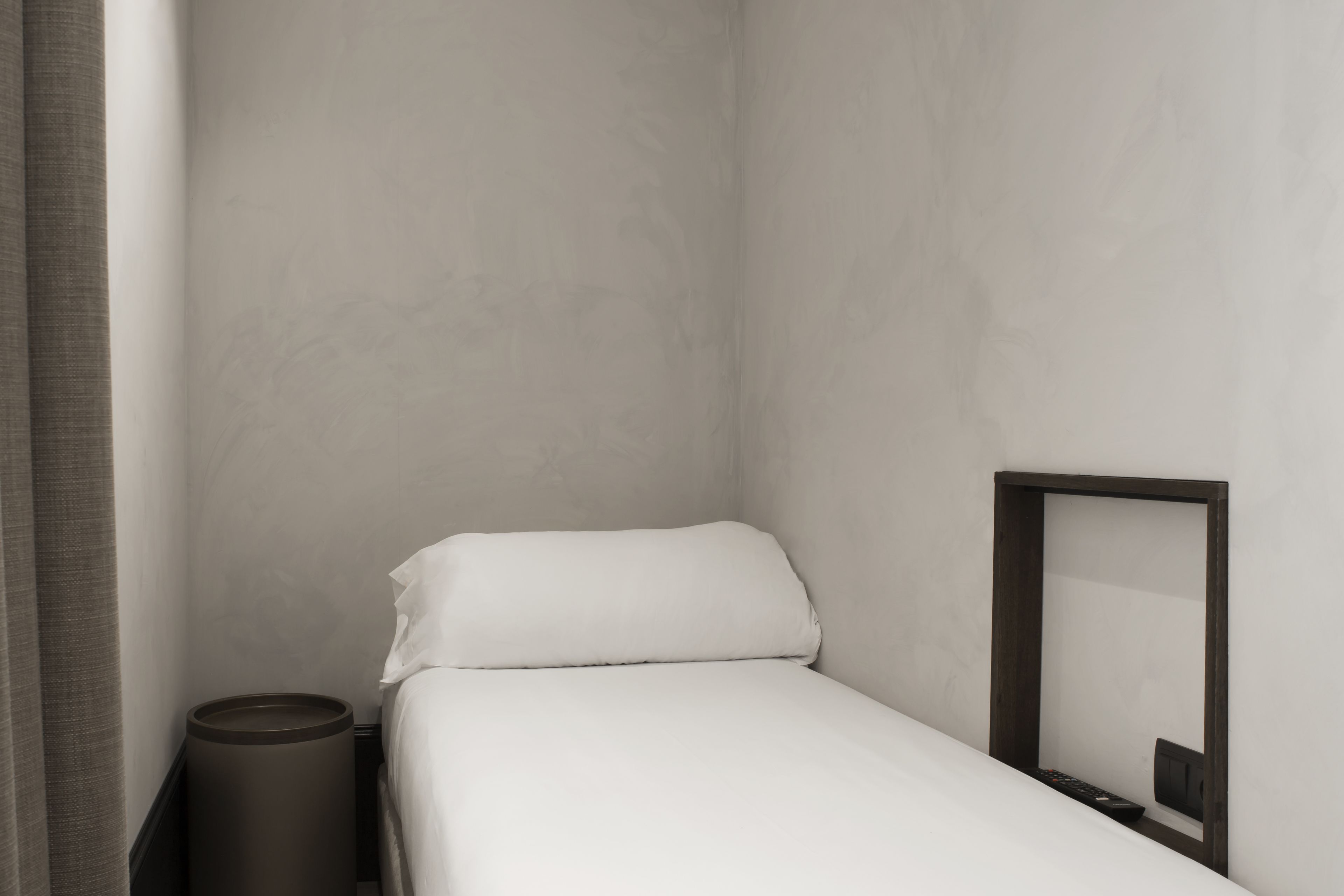 Una habitación pequeña con cama individual, pared sencilla, cubo de basura junto a la cama y un espejo.
