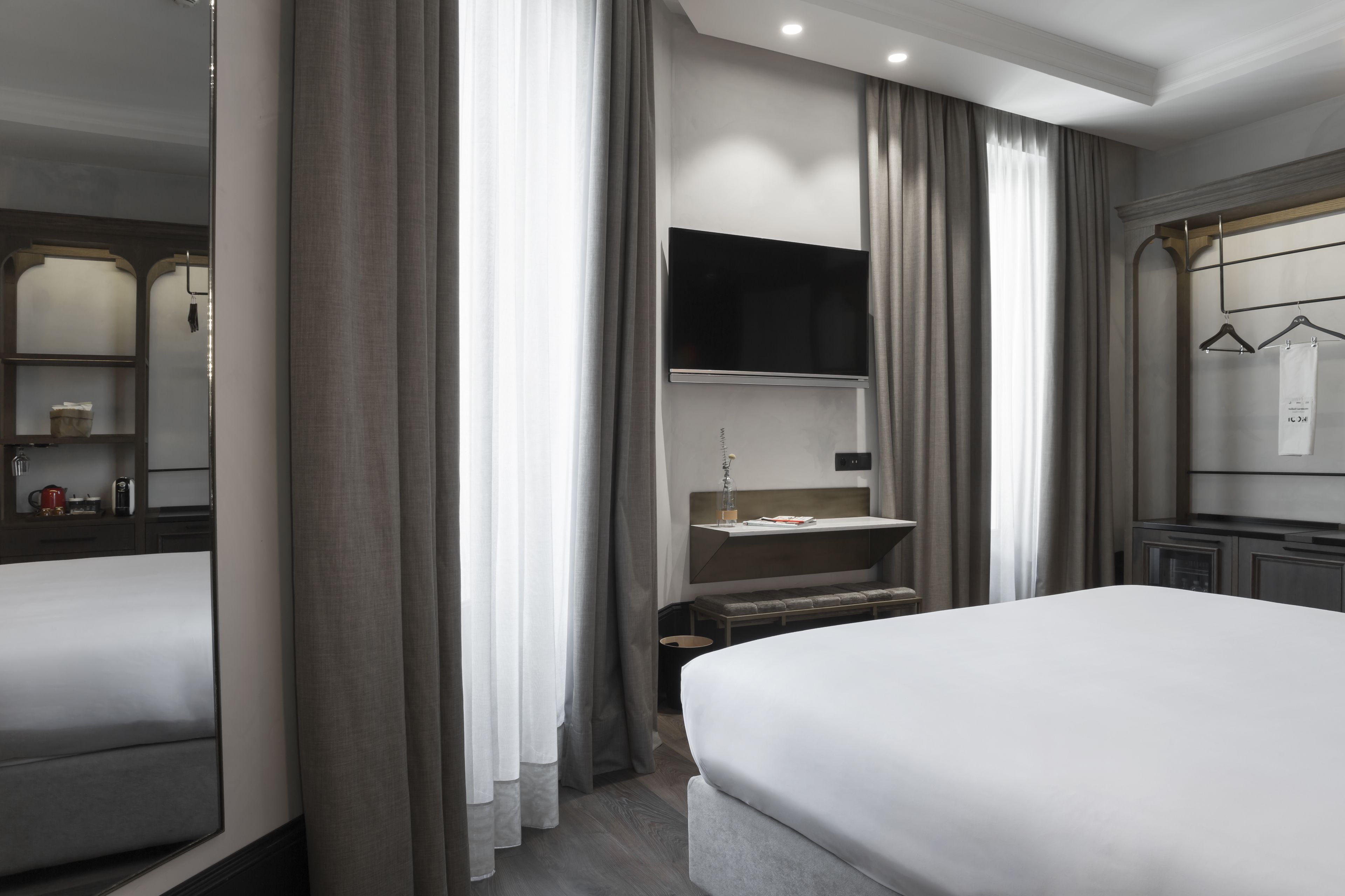 A modern hotel room with a large bed, a TV mounted on the wall, a small desk, and a bench.