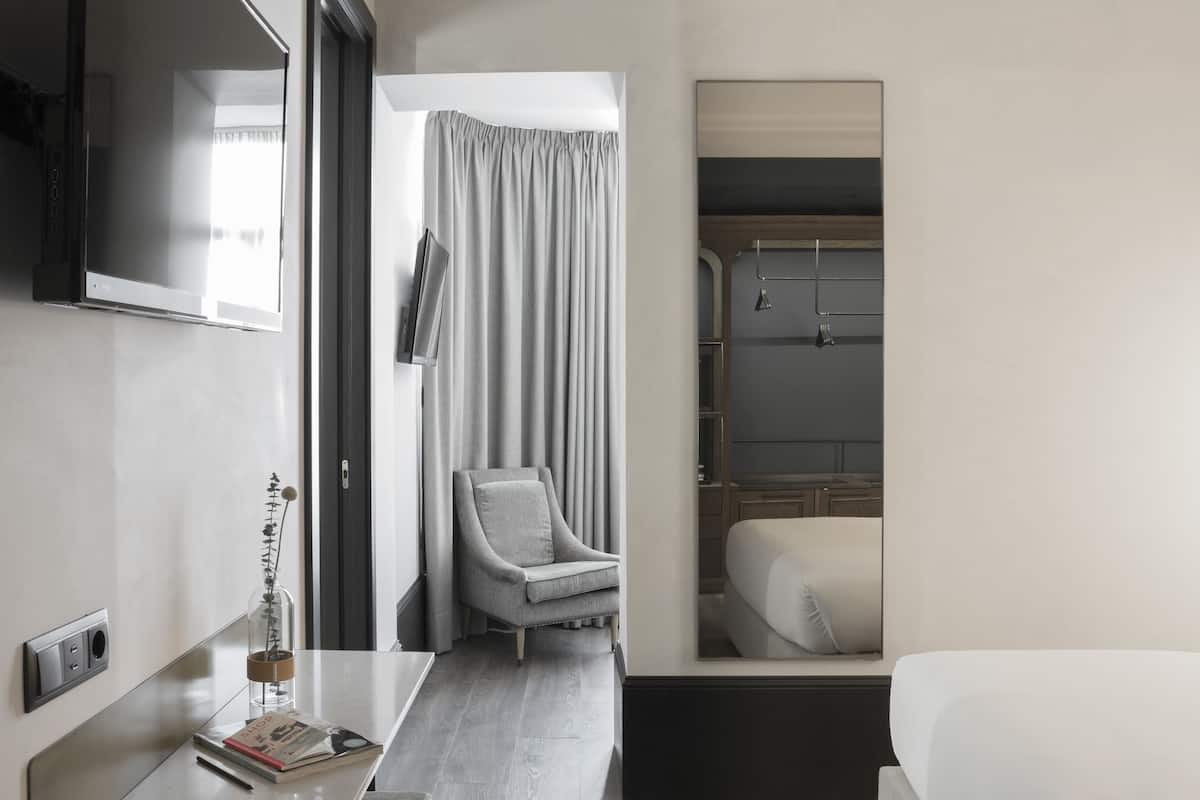A modern hotel room with a flat-screen TV mounted on the wall, a bedside table with a book and a decorative item, a grey armchair, a bed with white bedding, and a shower area visible through a glass door.