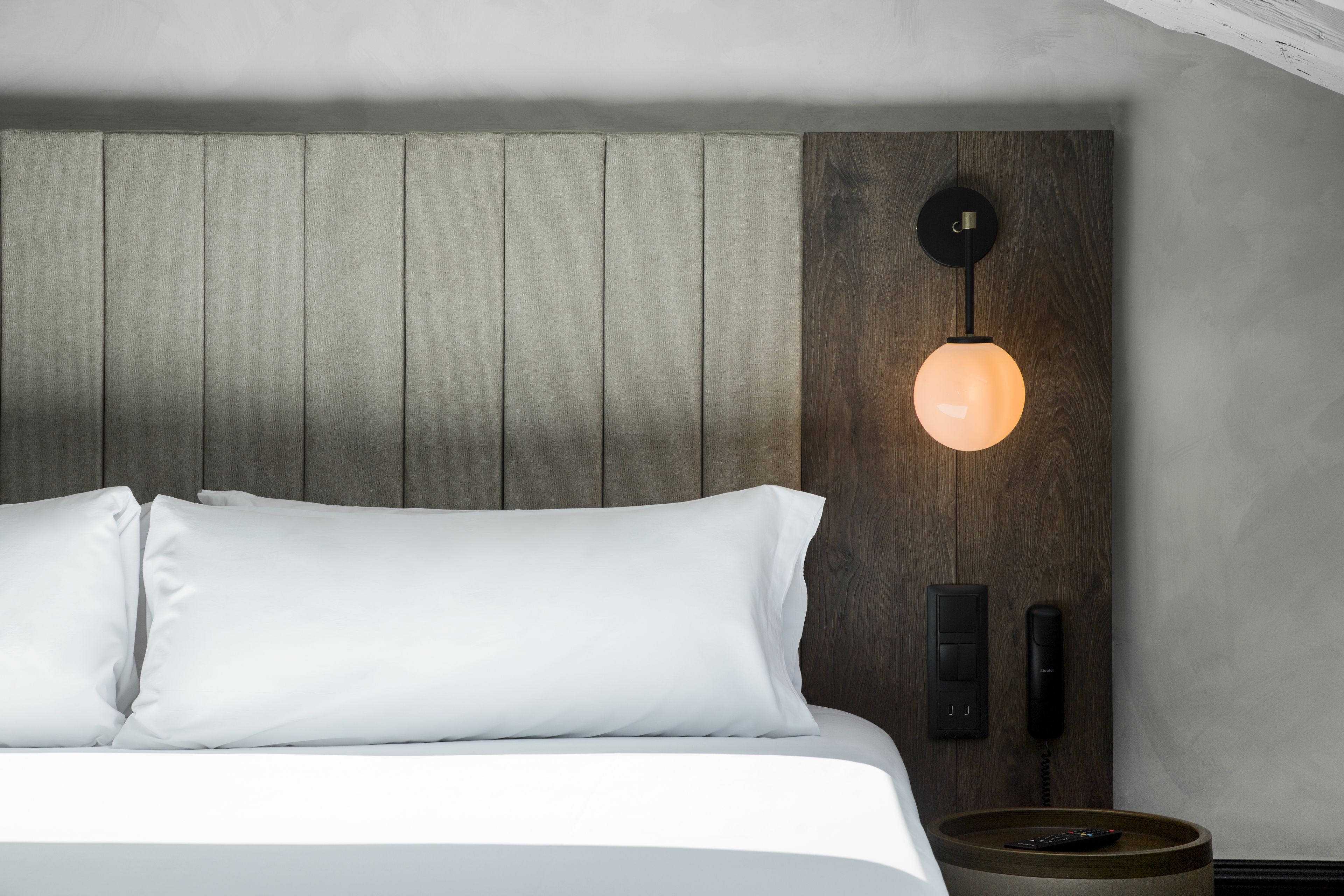 A modern hotel room with a bed, a wooden headboard, a wall-mounted phone, and a bedside table with a remote control.