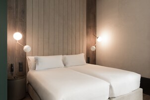 Ein Doppelbett mit weißer Bettwäsche, zwei Nachttischlampen, ein hölzernes Kopfteil und ein kleiner Nachttisch mit Telefon und Fernbedienung.