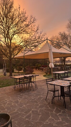 An outdoor seating area with tables and chairs under a canopy, surrounded by trees and a sunset in the background.