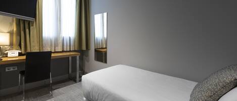 Una habitación de hotel con cama, escritorio, silla, lámpara y ventana con cortinas.
