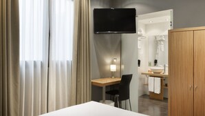 Habitació d'hotel moderna amb televisió de pantalla plana penjada a la paret, escriptori amb cadira, armari de fusta i bany amb mirall i rentapelats.